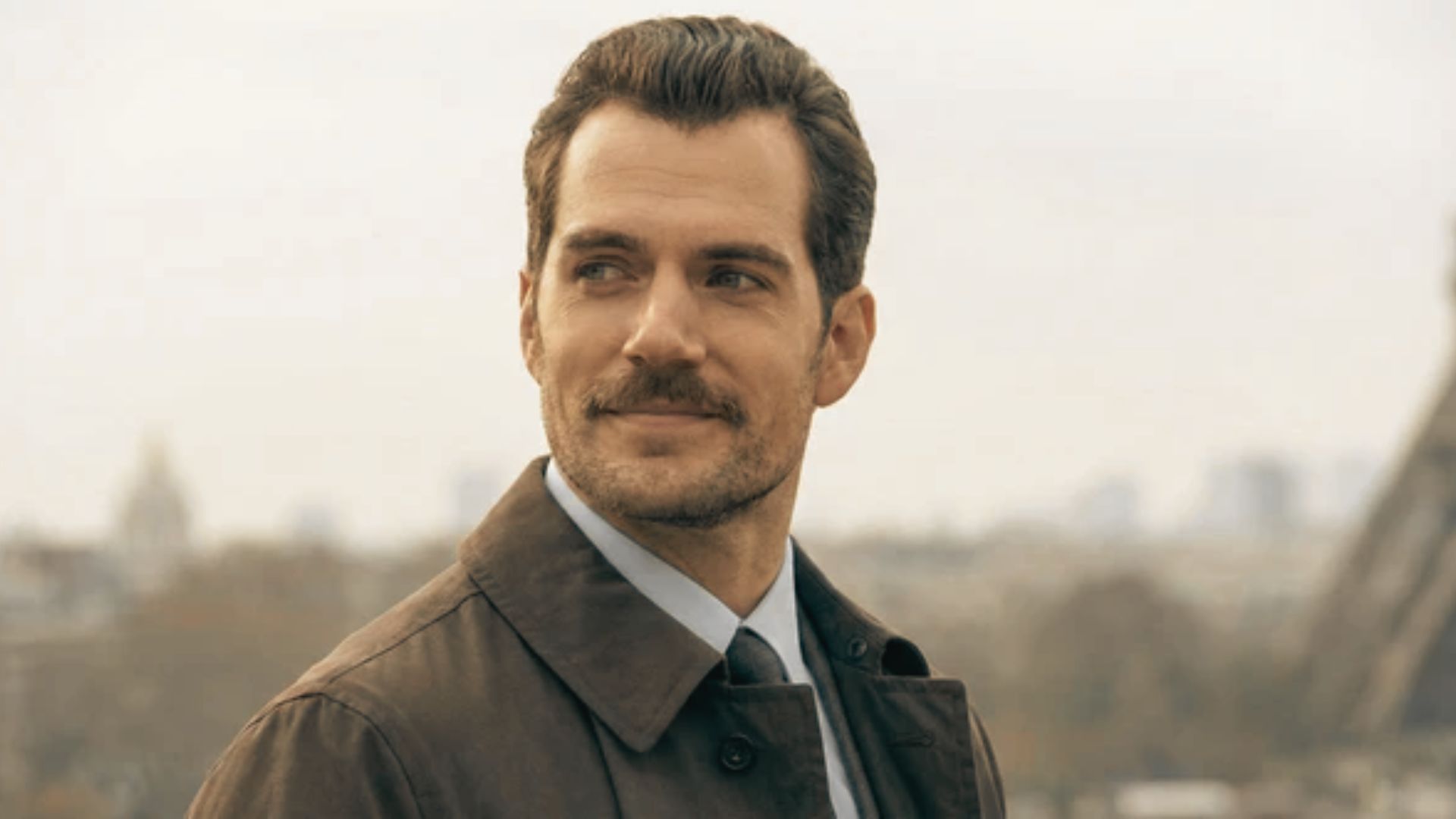 Henry Cavill in a brown coat and tie stands confidently in front of the Eiffel Tower, showcasing a slicked-back hairstyle.