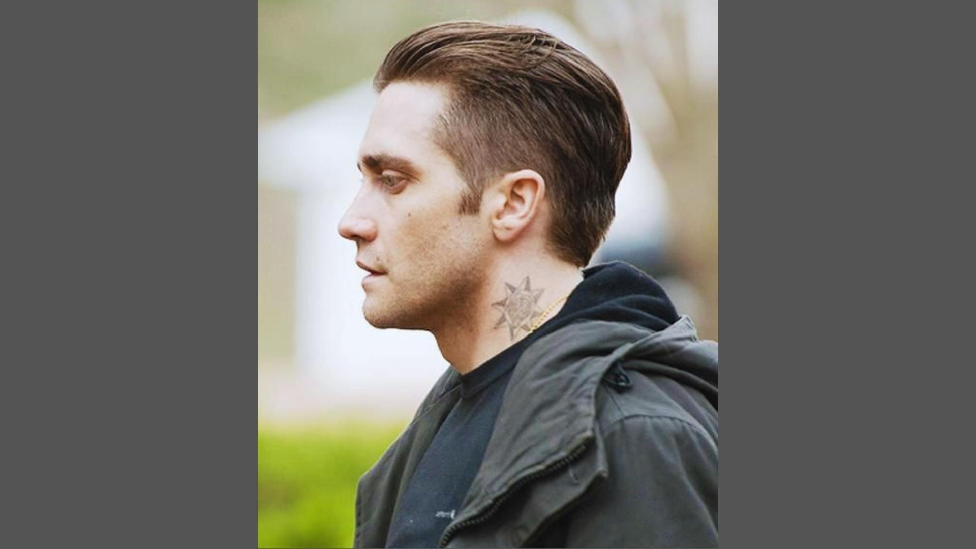 Side profile of Jake Gyllenhaal with modern slicked-back undercut haircut, tapered sides, and neck tattoo wearing black jacket.