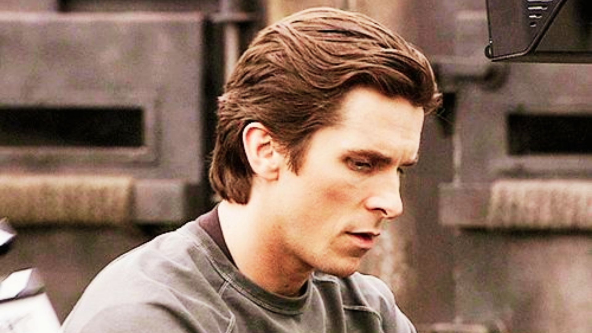 A man in a grey shirt gazes intently, showcasing a hairstyle reminiscent of Christian Bale in The Dark Knight Rises.