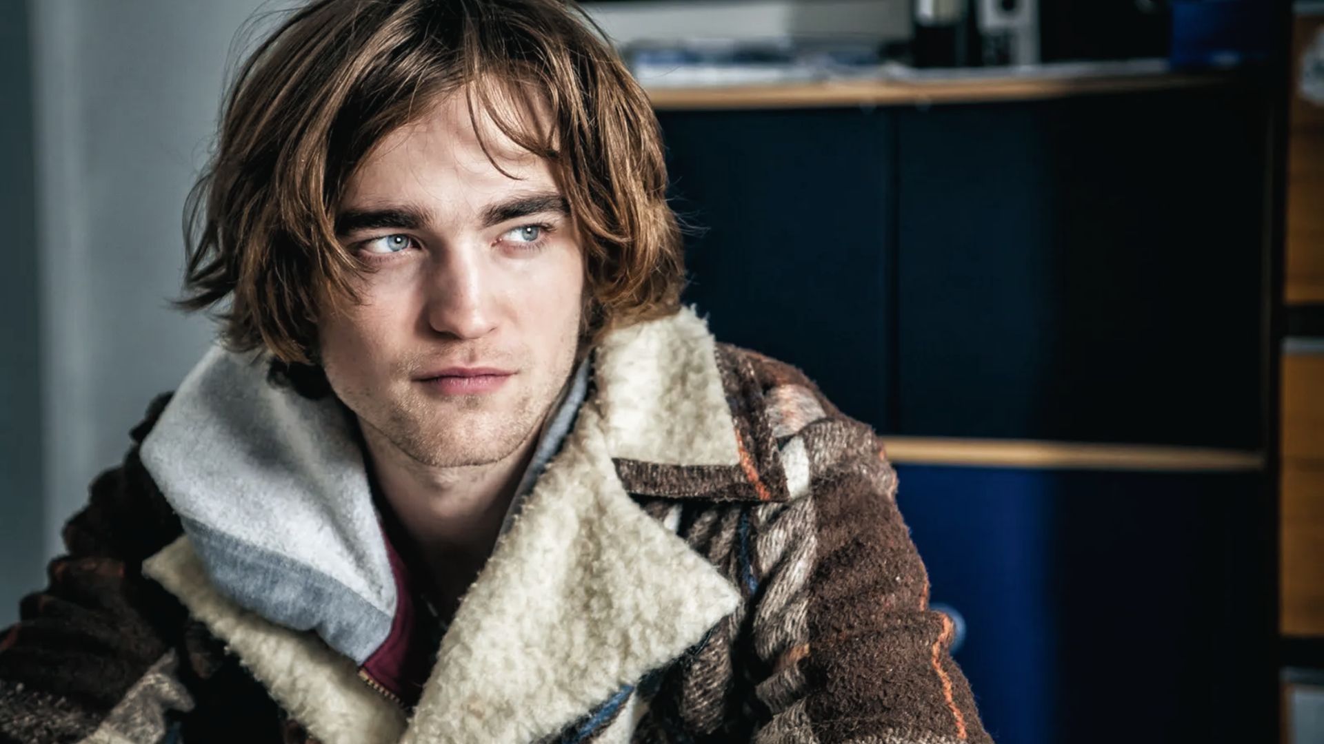 Robert Pattinson in a jacket sitting at a table, showcasing long hair style features.