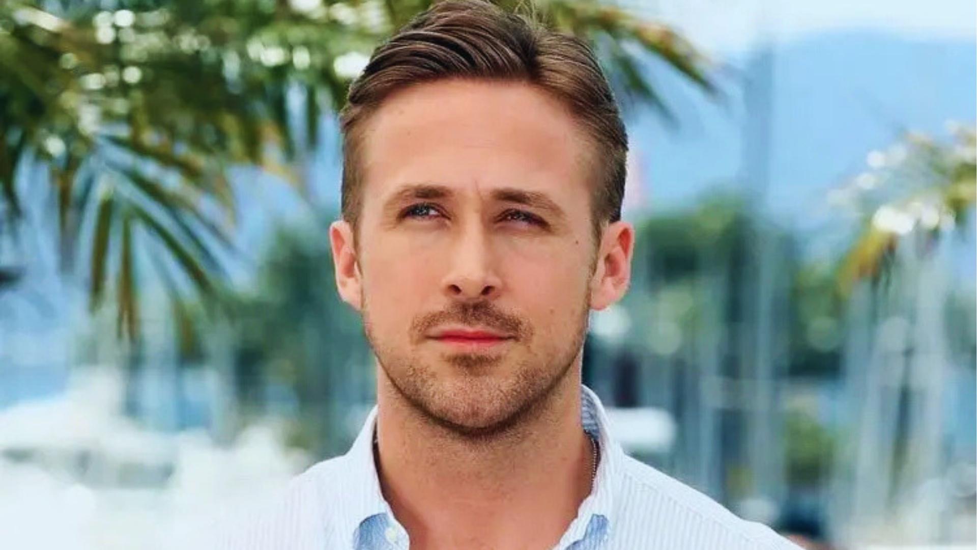 Ryan Gosling with short textured comb-over haircut, light stubble, and tapered sides wearing light blue striped shirt against palm backdrop.