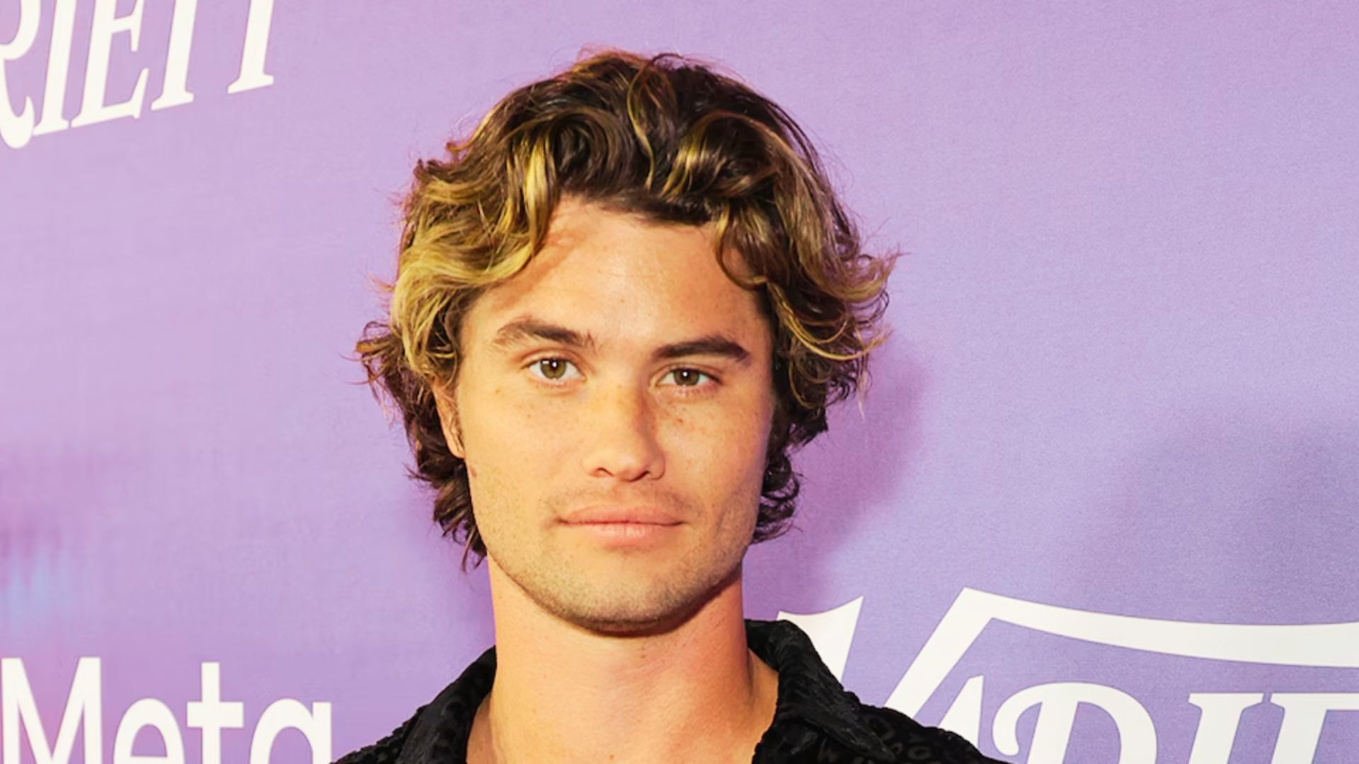 A man with curly hair wearing a black shirt, contemplating the suitability of the John B haircut for himself.