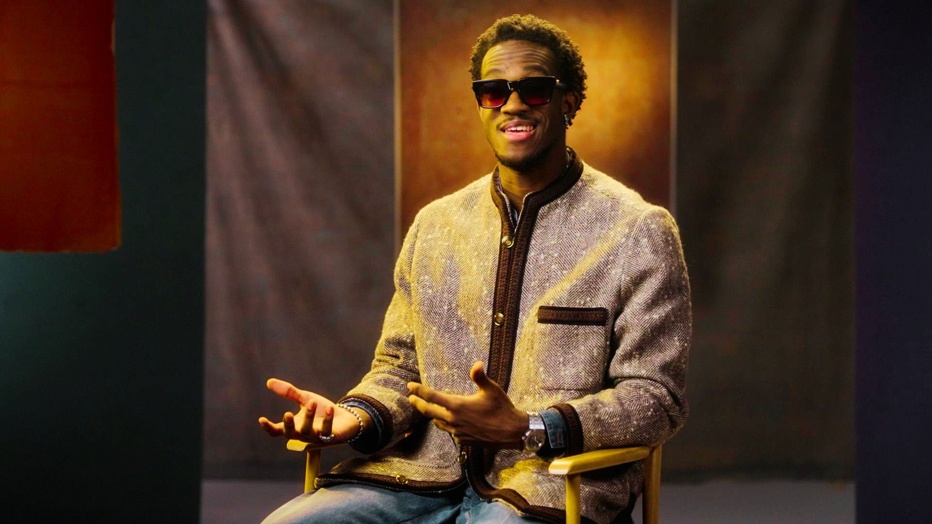 A man in sunglasses sits on a chair, hands outstretched, showcasing a stylish outerwear outfit.