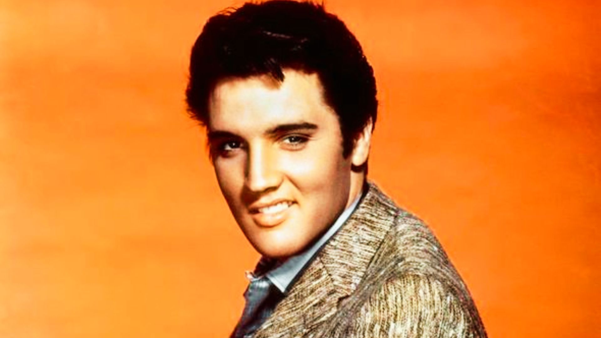 Elvis Presley poses against an orange background, showcasing his iconic hairstyle.