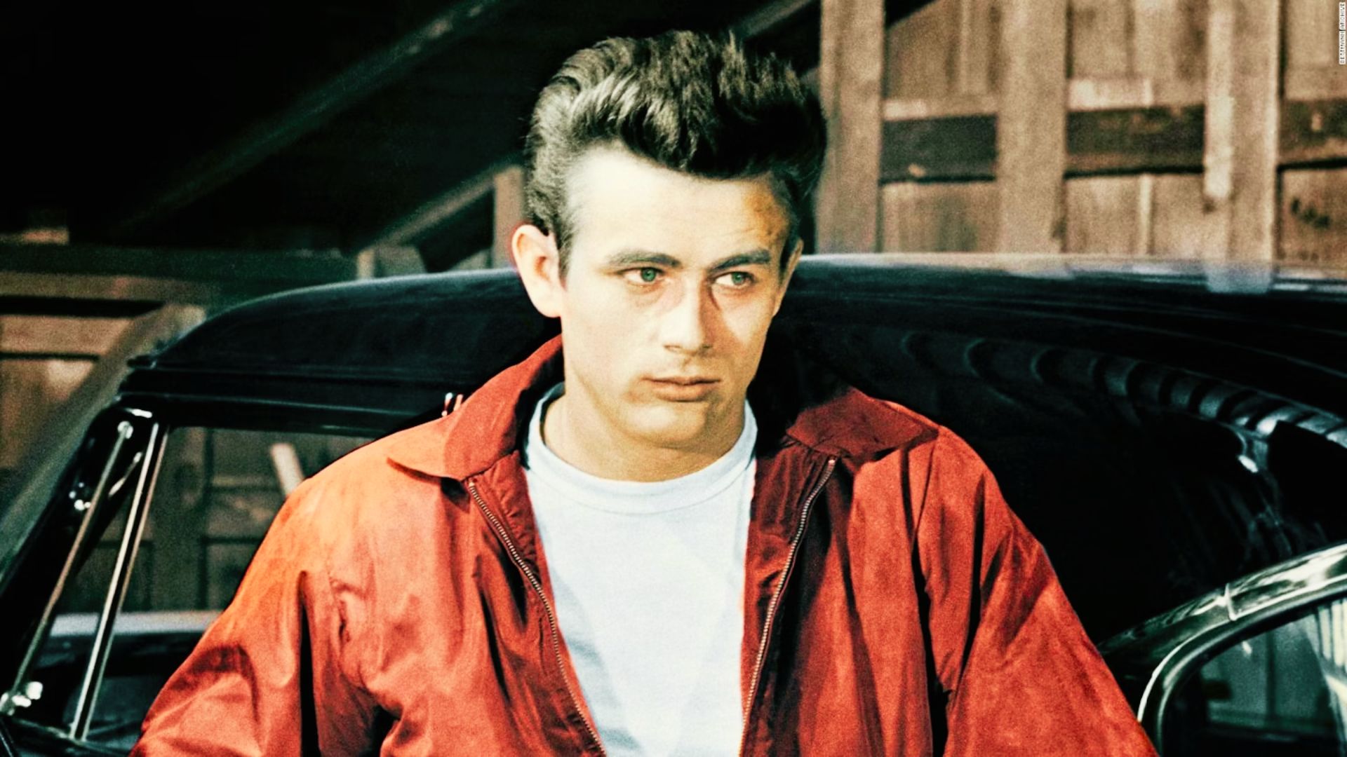 James Dean in a red jacket stands beside a black car, showcasing a modern hairstyle.