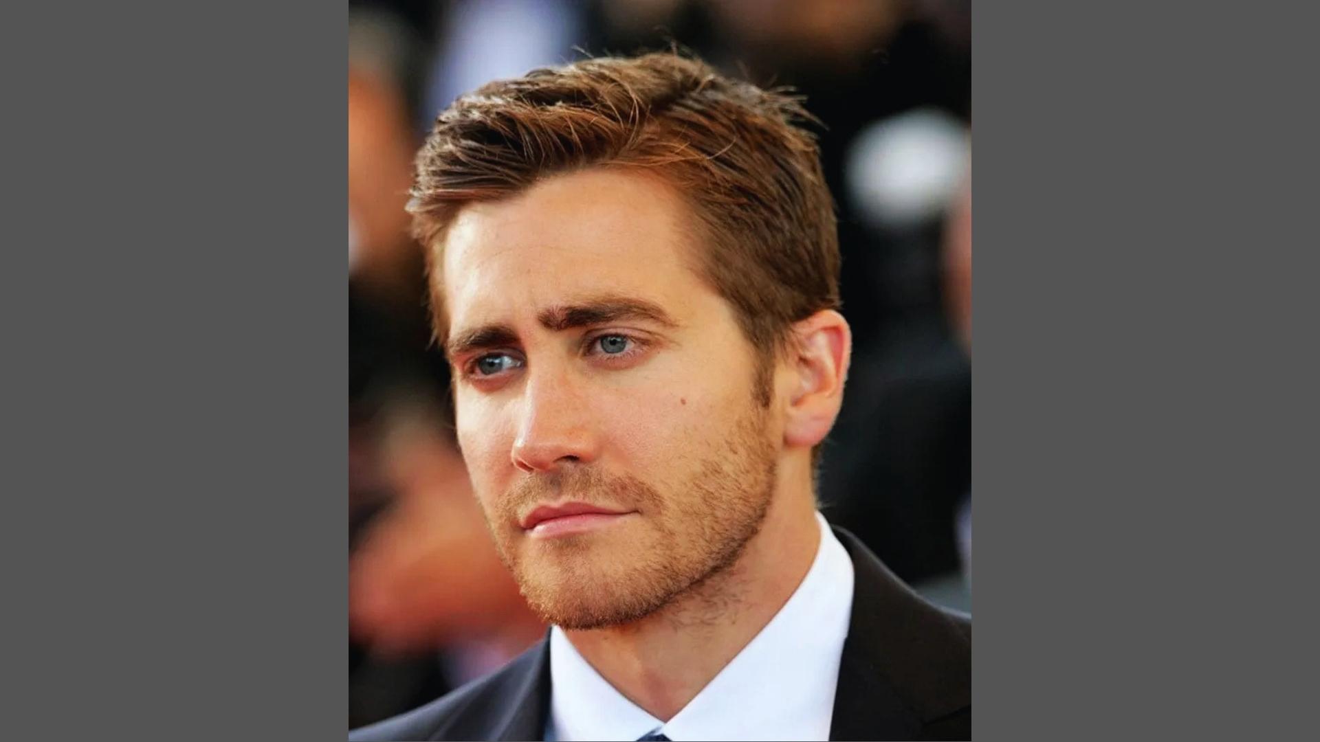 Jake Gyllenhaal with classic side-swept haircut, textured top, and light stubble wearing suit at red carpet.