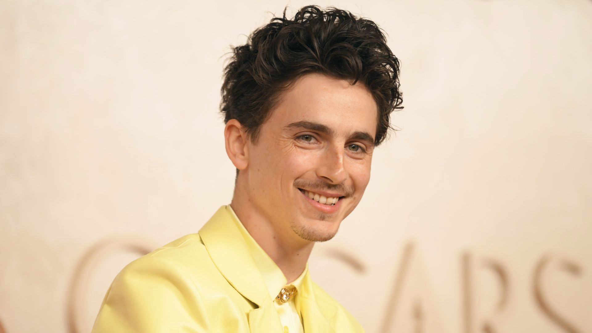 Timothee Chalamet in curly hair dressed in a bright yellow suit, standing confidently with a neutral expression.