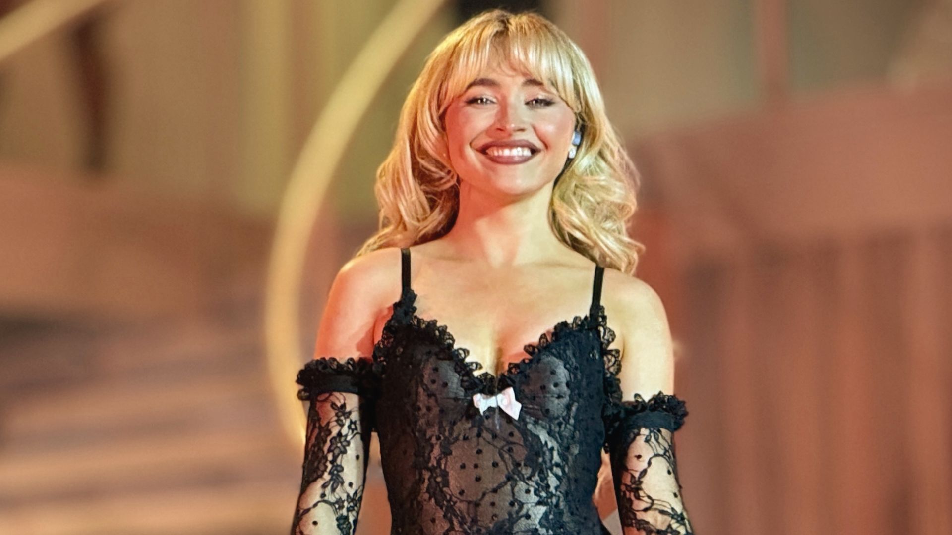 Sabrina Carpenter smiles on stage in a black sheer lace top with matching long gloves and a white bow.