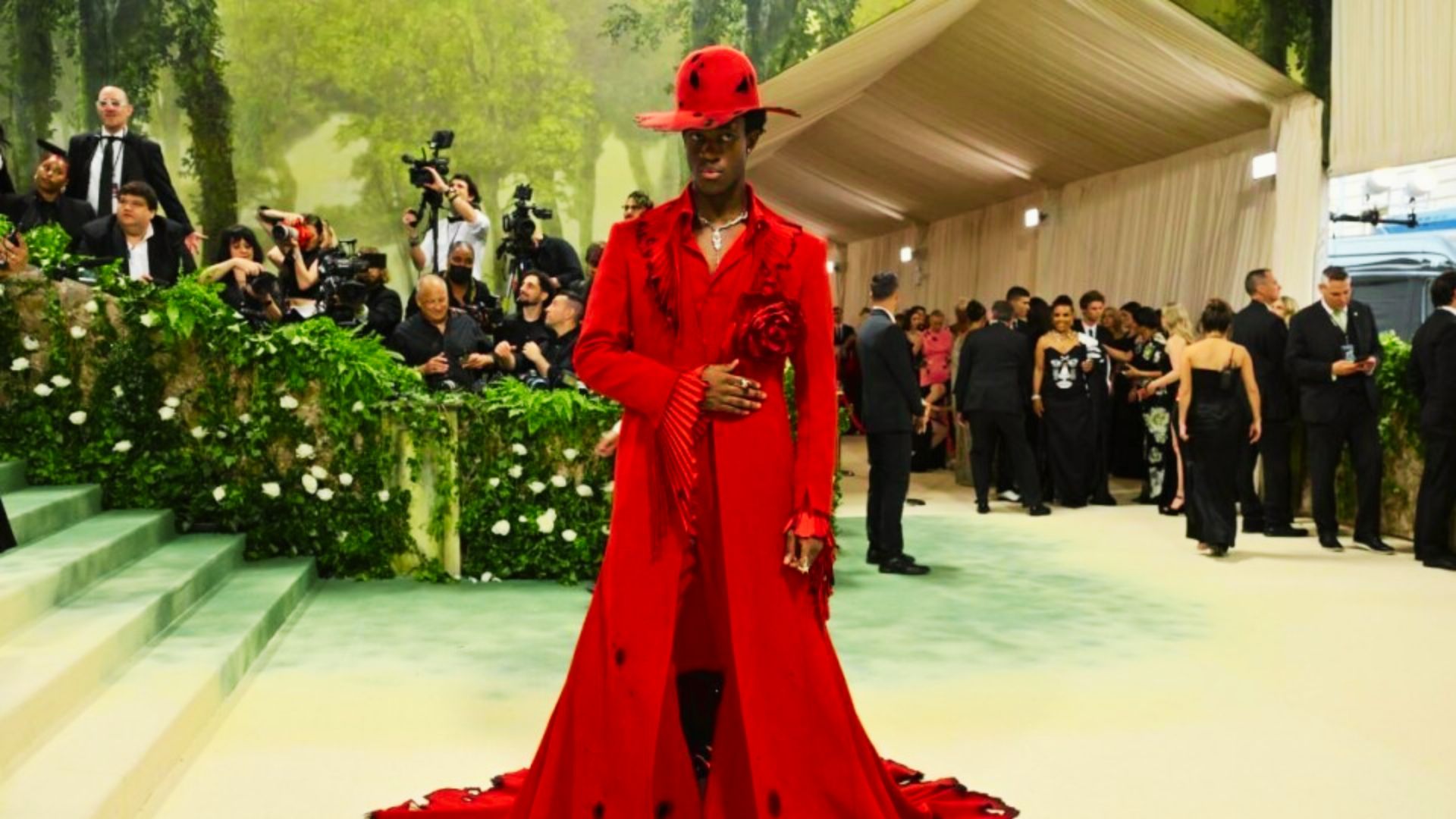Wisdom Kaye in a red dress and hat stands properly on a carpet