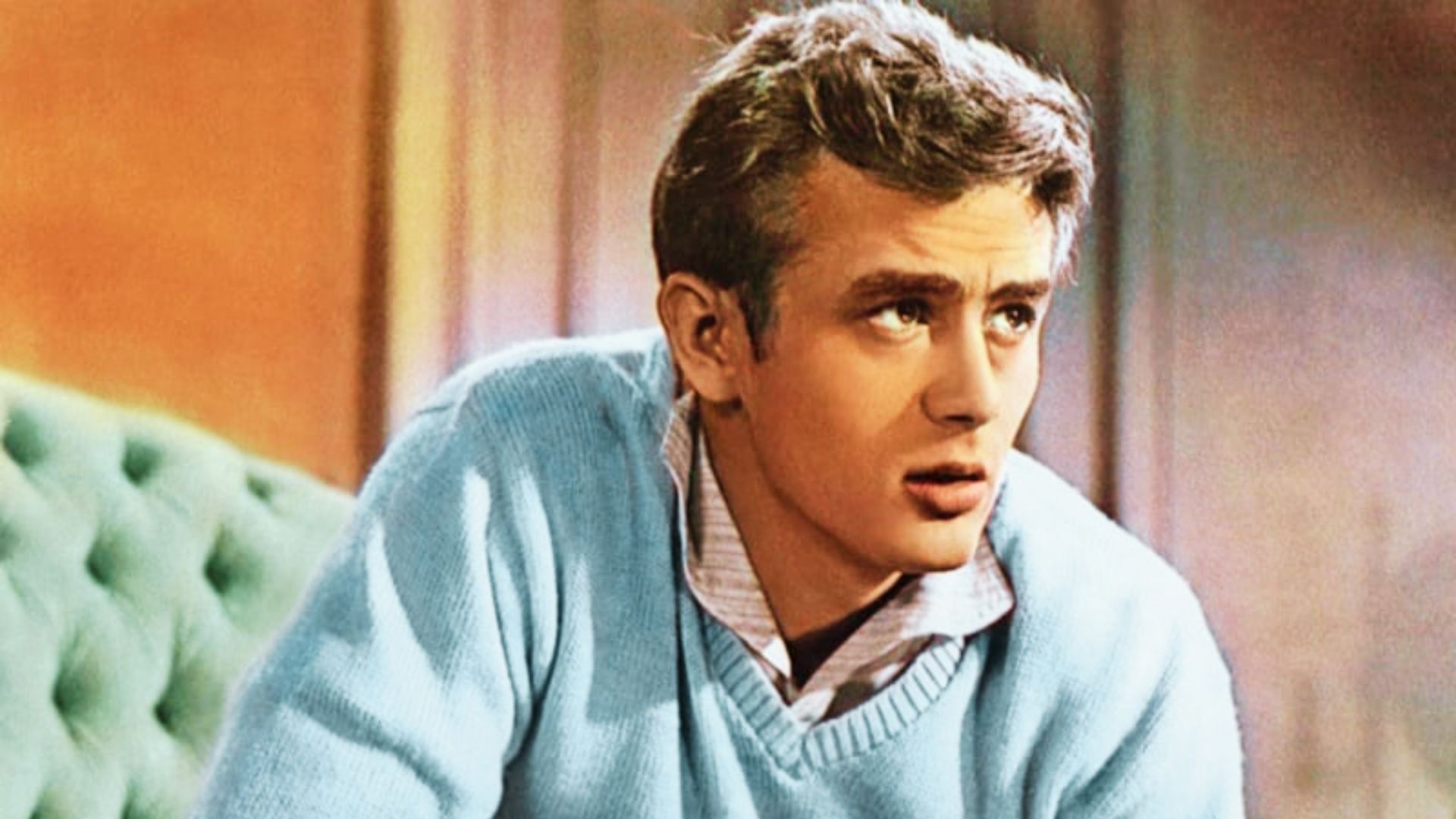James Dean in a blue sweater sits on a couch, hairstyle for a timeless look.