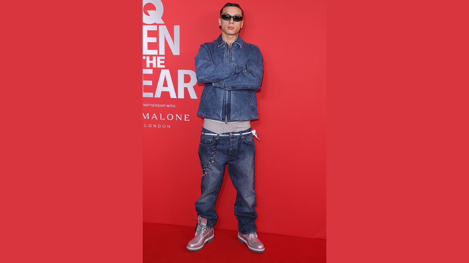 "A man wearing jeans and a jacket stands confidently on a vibrant red carpet."