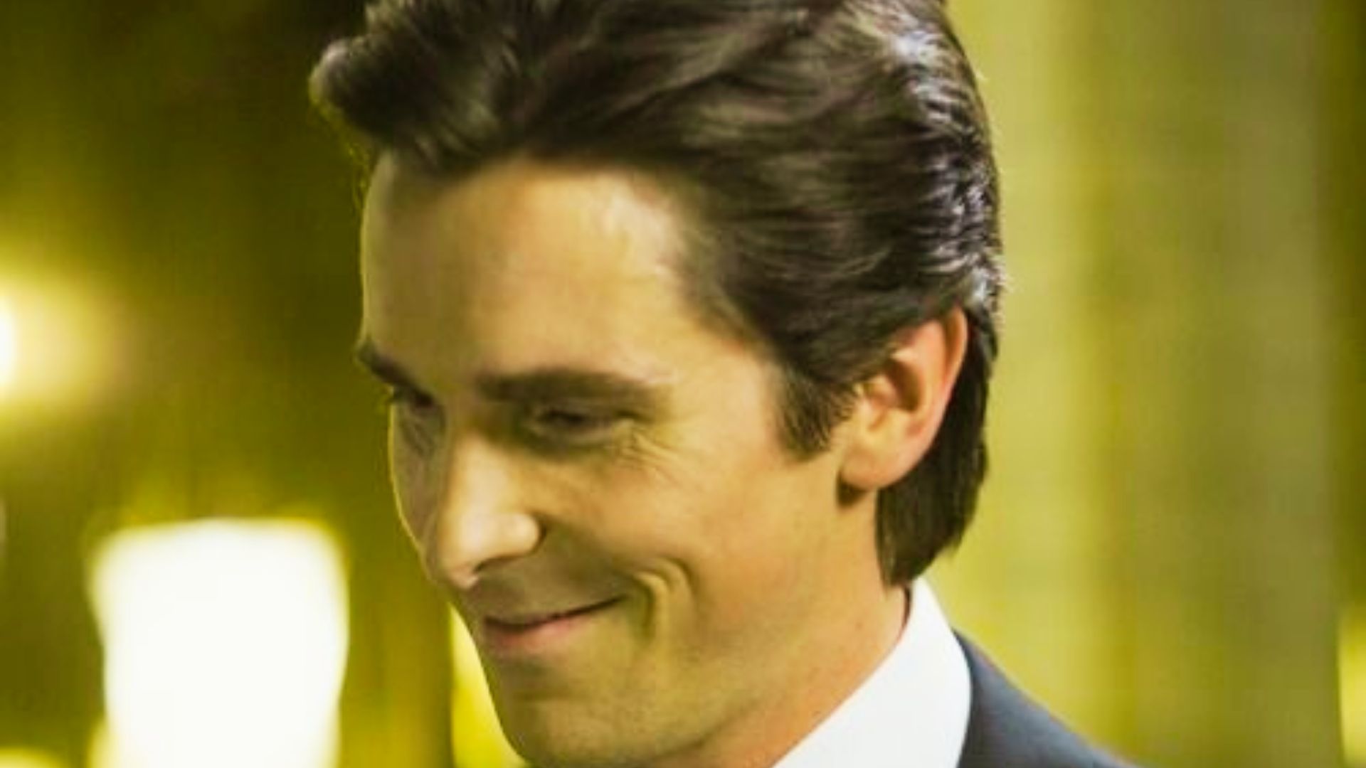 Christian Bale' in a suit and tie, smiling, with a hairstyle in The Dark Knight.