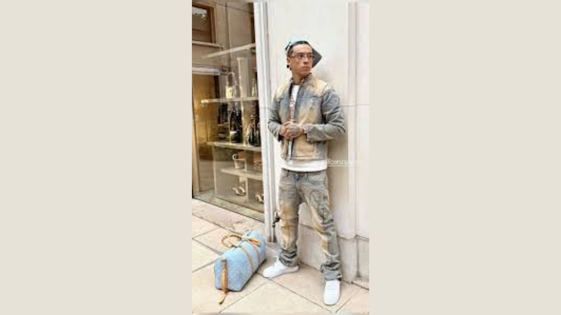 "A man wearing jeans and a jacket stands next to a blue bag, looking casually toward the camera."