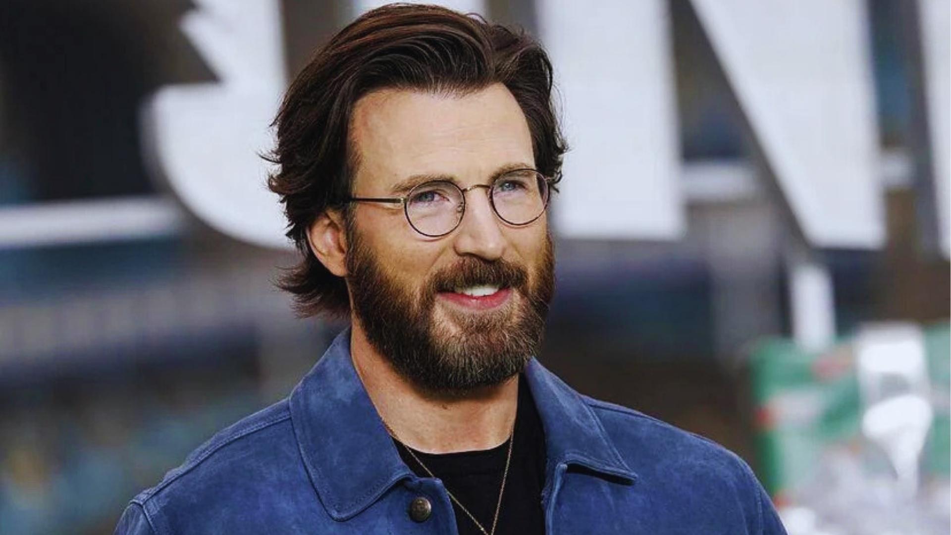 Chris Evans with long layered flow haircut reaching collar, middle part, round glasses, and full beard in blue suede jacket.