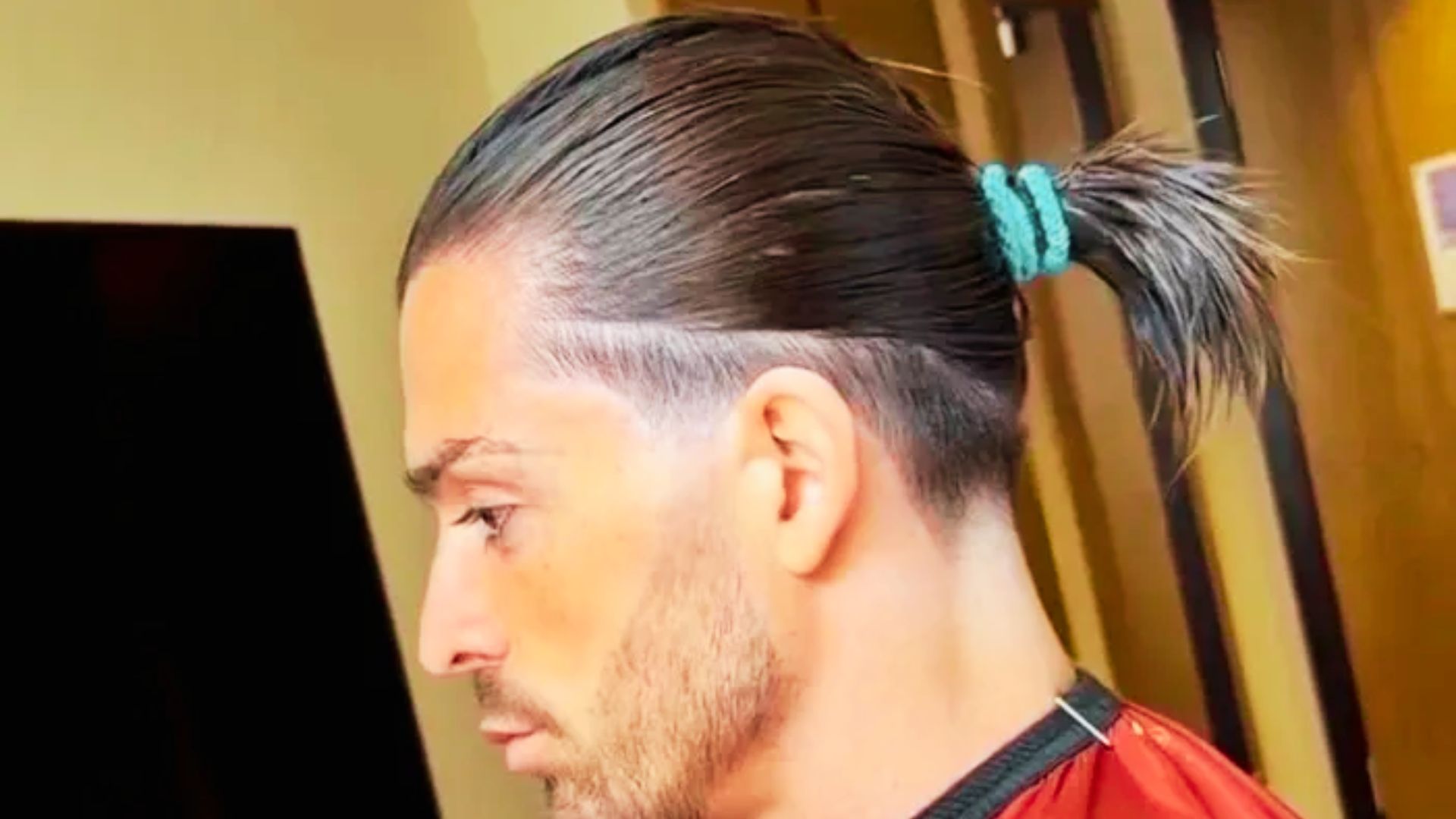 Jack Grealish with a ponytail sits in a barber shop, preparing for an undercut as part of a haircut tutorial.