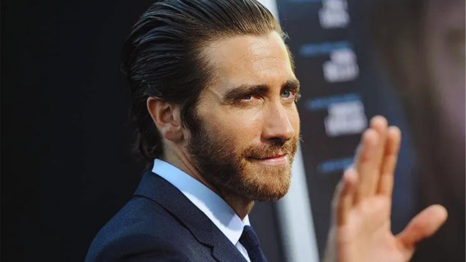 Jake Gyllenhaal with slicked-back long haircut and full beard waving while wearing dark suit and tie at premiere.