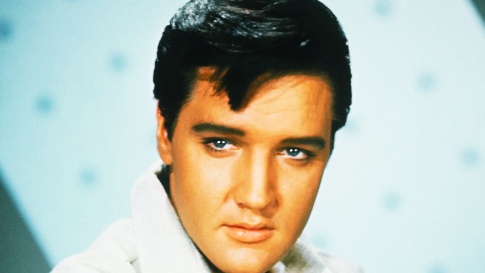 Elvis Presley wearing a white shirt, showcasing his blue eyes and messy voluminous hairstyle.