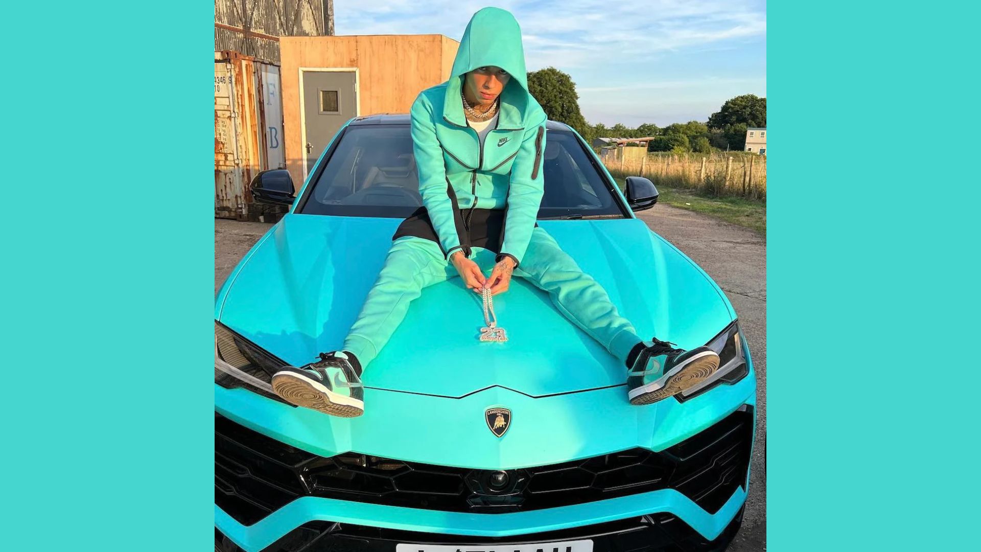 "A man wearing a hoodie sits on the hood of a blue car, appearing relaxed and casual."