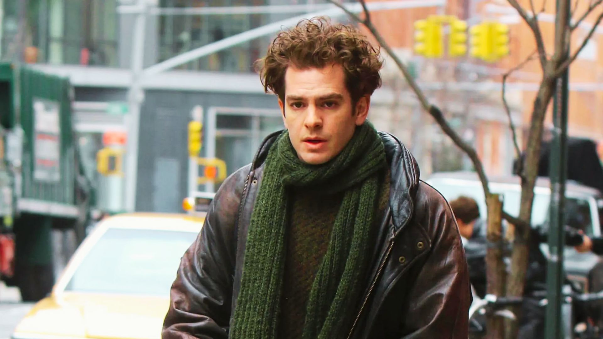 Andrew Garfield in a brown jacket and green scarf walks down the street