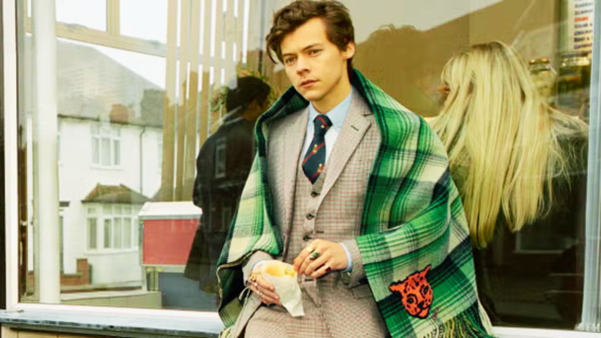 Harry Styles sits by a window in a plaid three-piece suit draped with a green tartan blanket.
