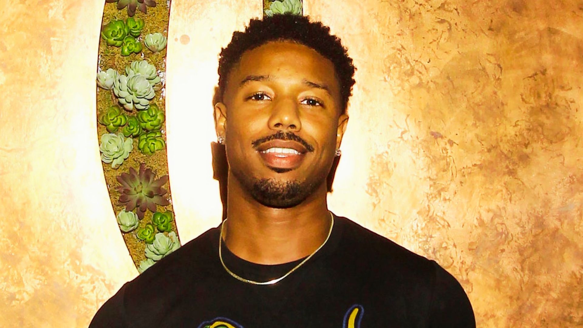 Michael B. Jordan smiling in a black shirt, wearing a short curly Afro with a rounded hairline.