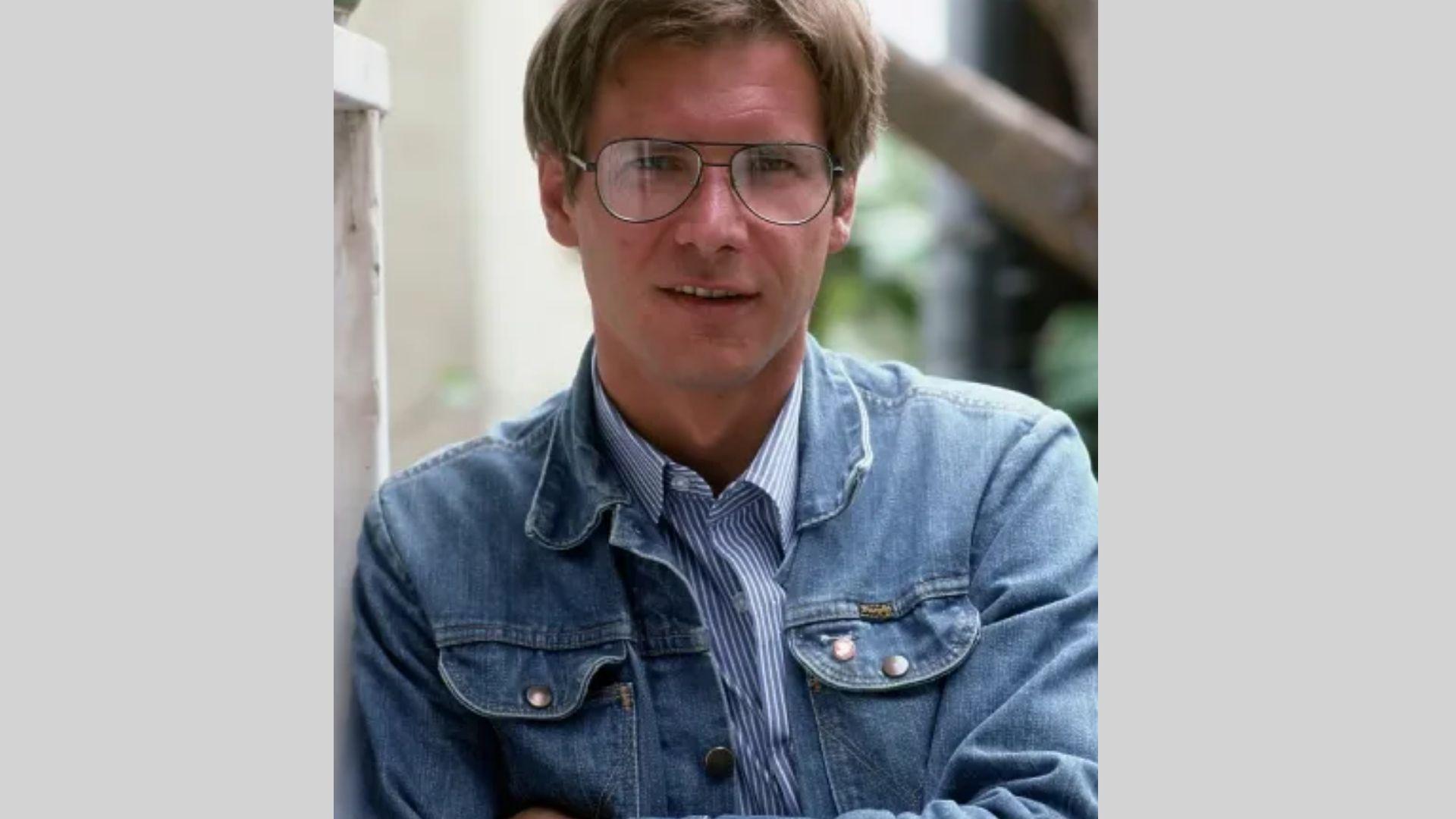 Harrison Ford in denim jacket and striped shirt wears glasses leaning against white wall