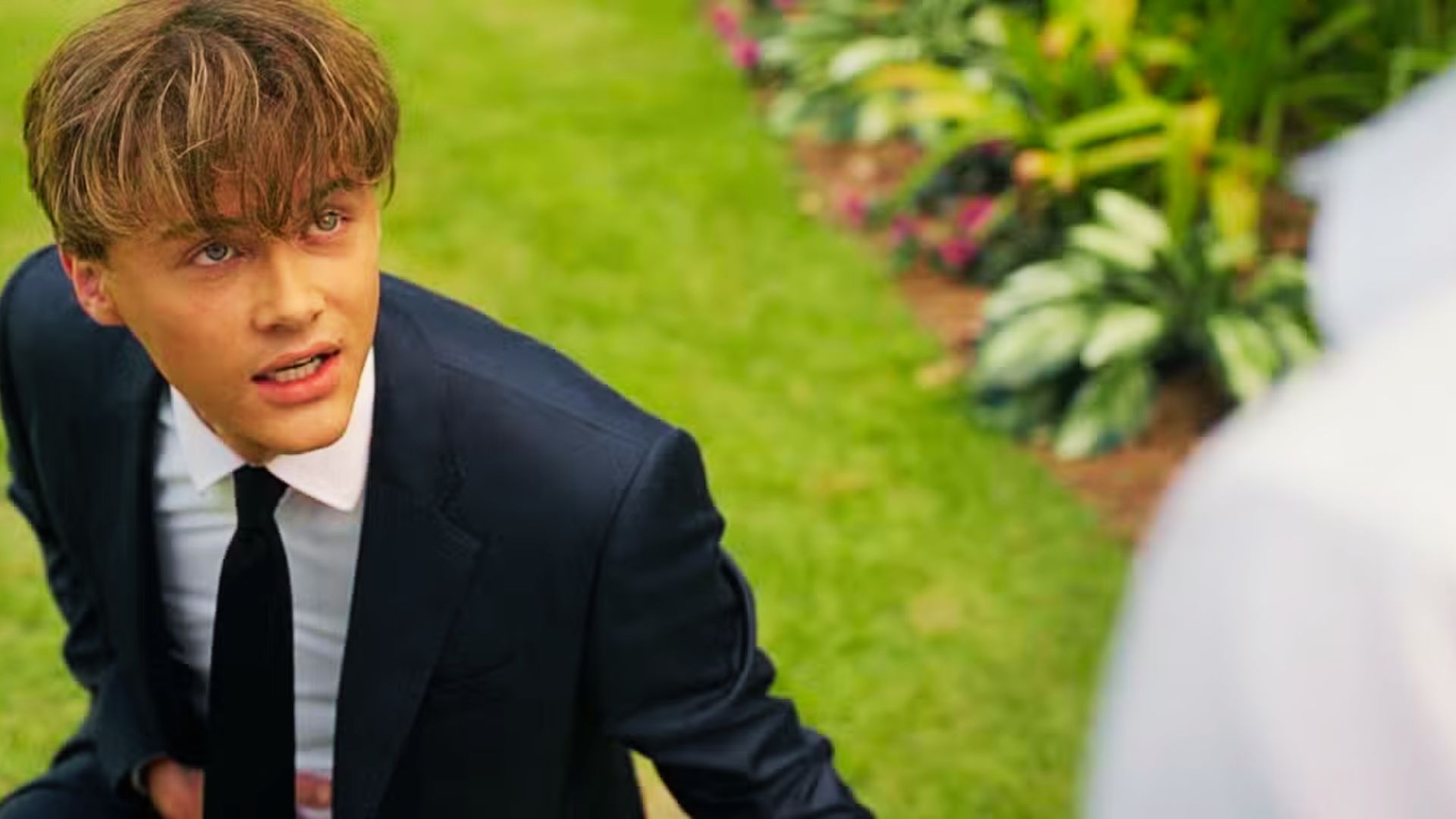 Conrad kneels on grass in a sharp black suit with a white shirt and black tie.