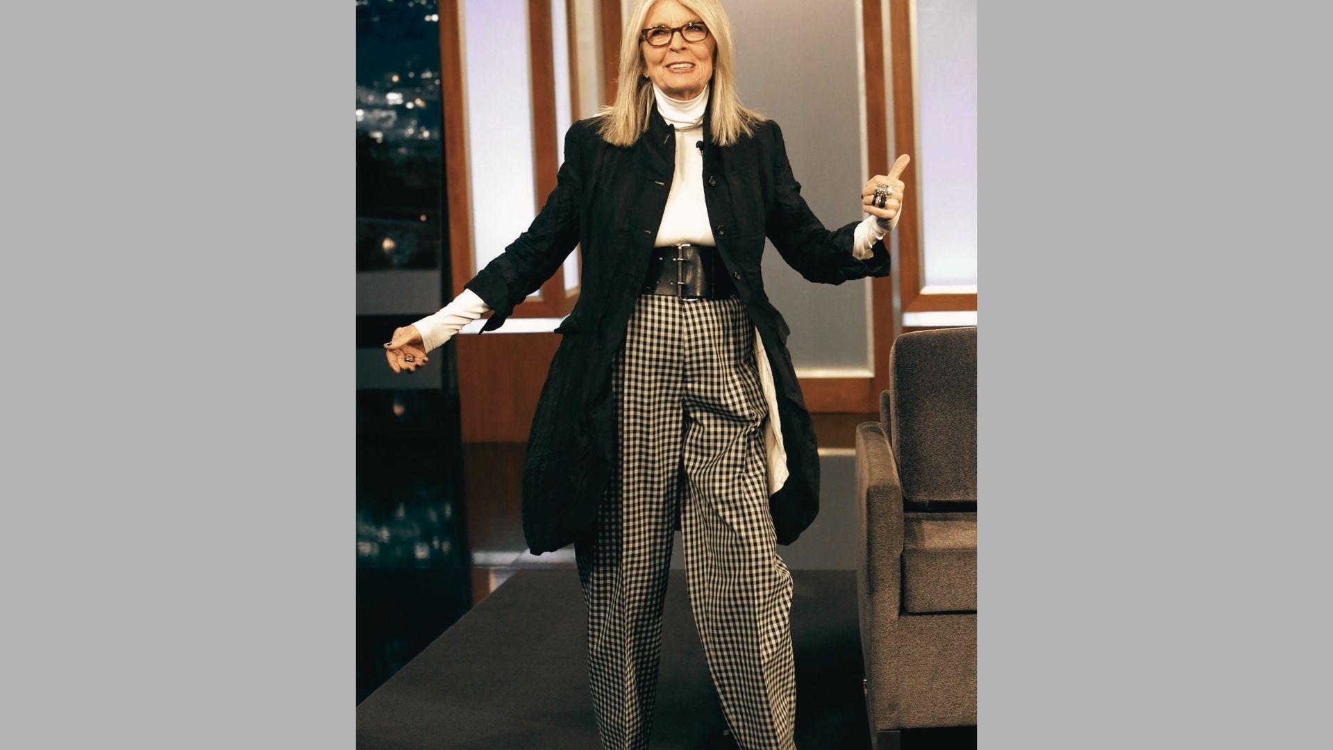 Woman in black jacket, white turtleneck, and checkered pants gestures on talk show set