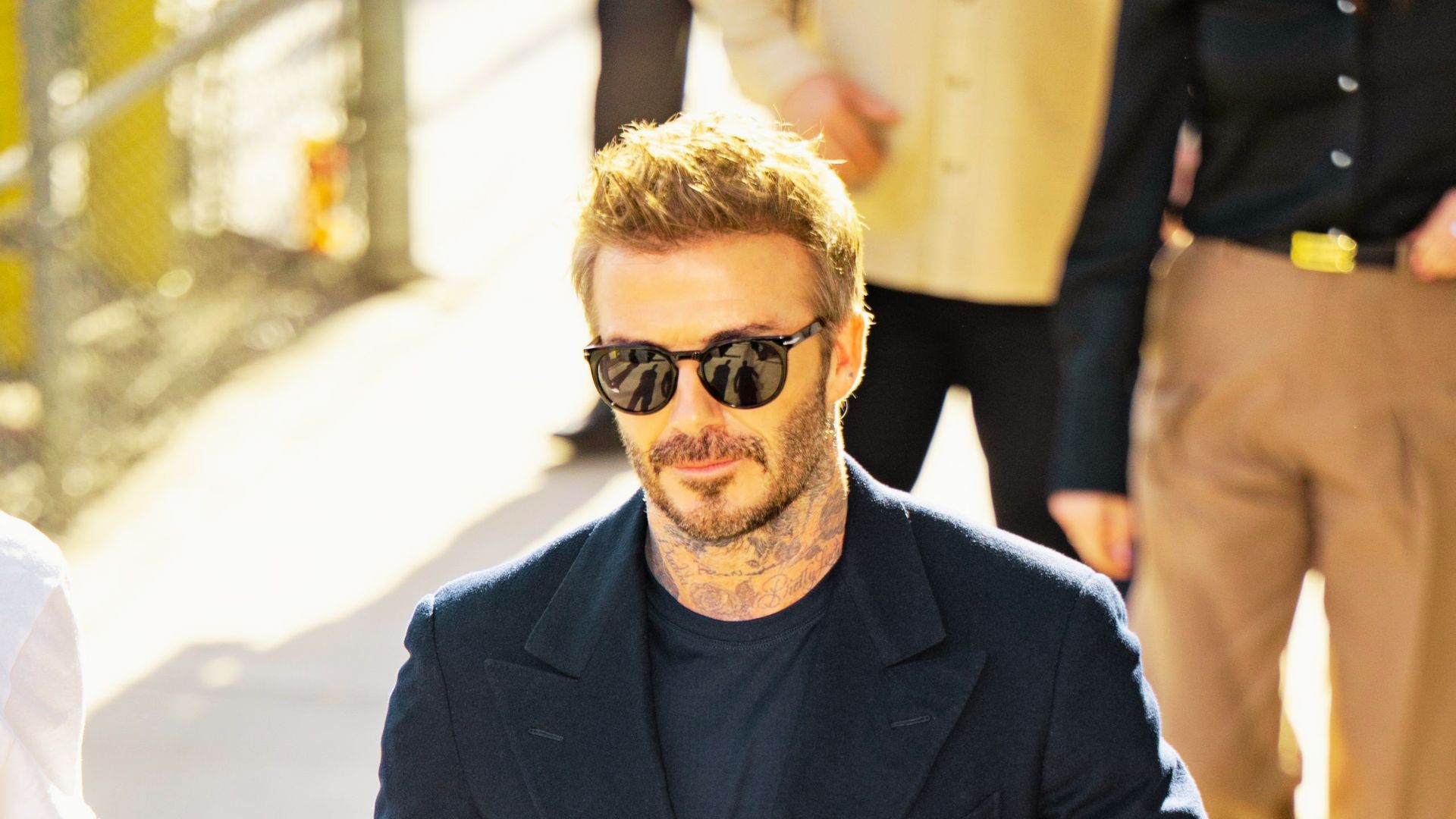 David Beckham walks in sunlight wearing a tailored navy blazer over a black tee and round sunglasses.