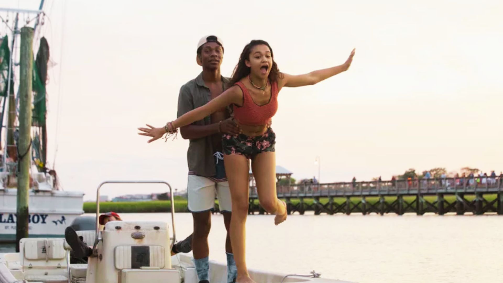 Kiara balances playfully on a boat in a red cropped tank top and black floral shorts, arms outstretched.