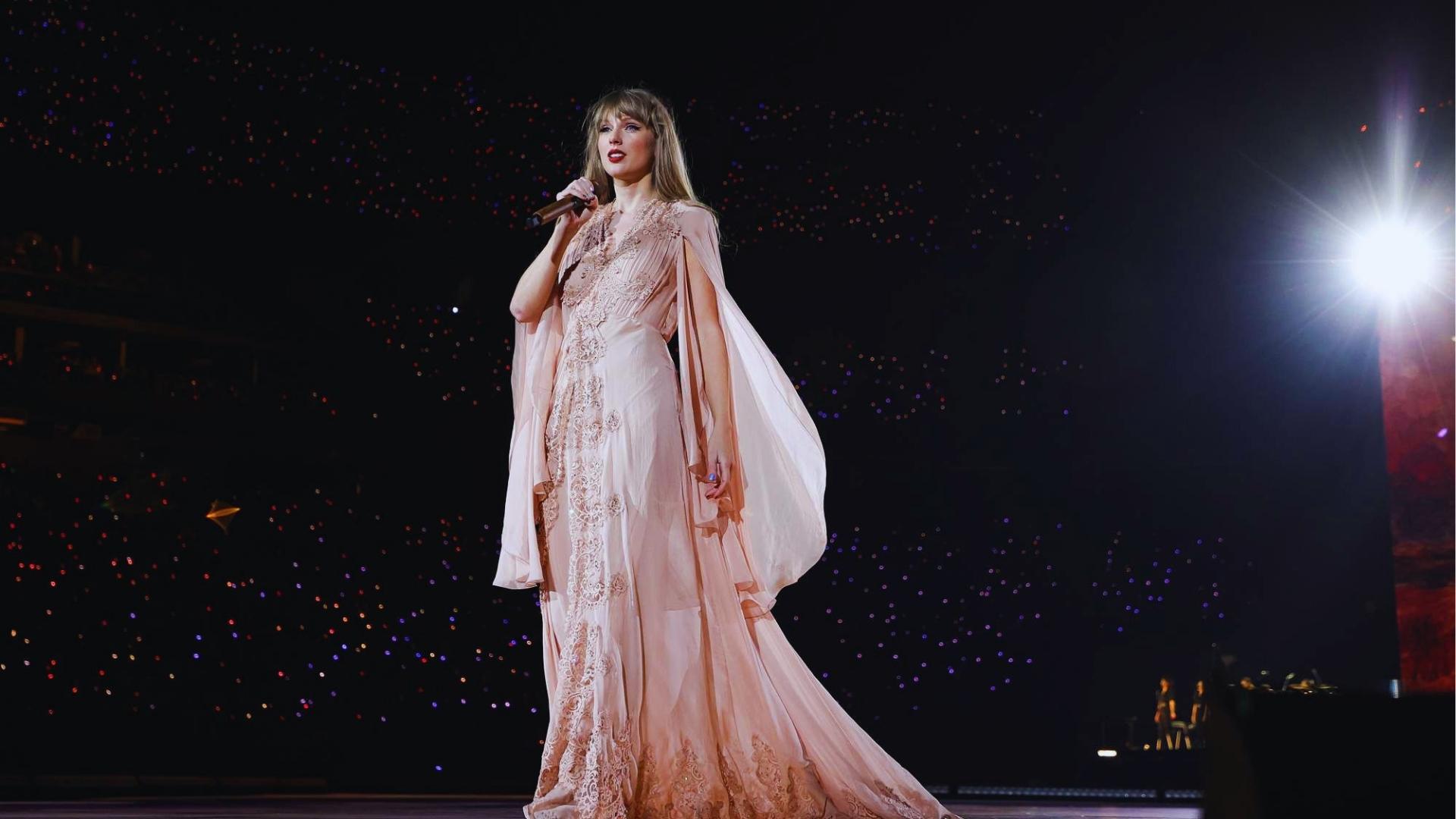 Taylor Swift performs in a blush pink lace folklore gown with sheer cape sleeves against stadium lights.