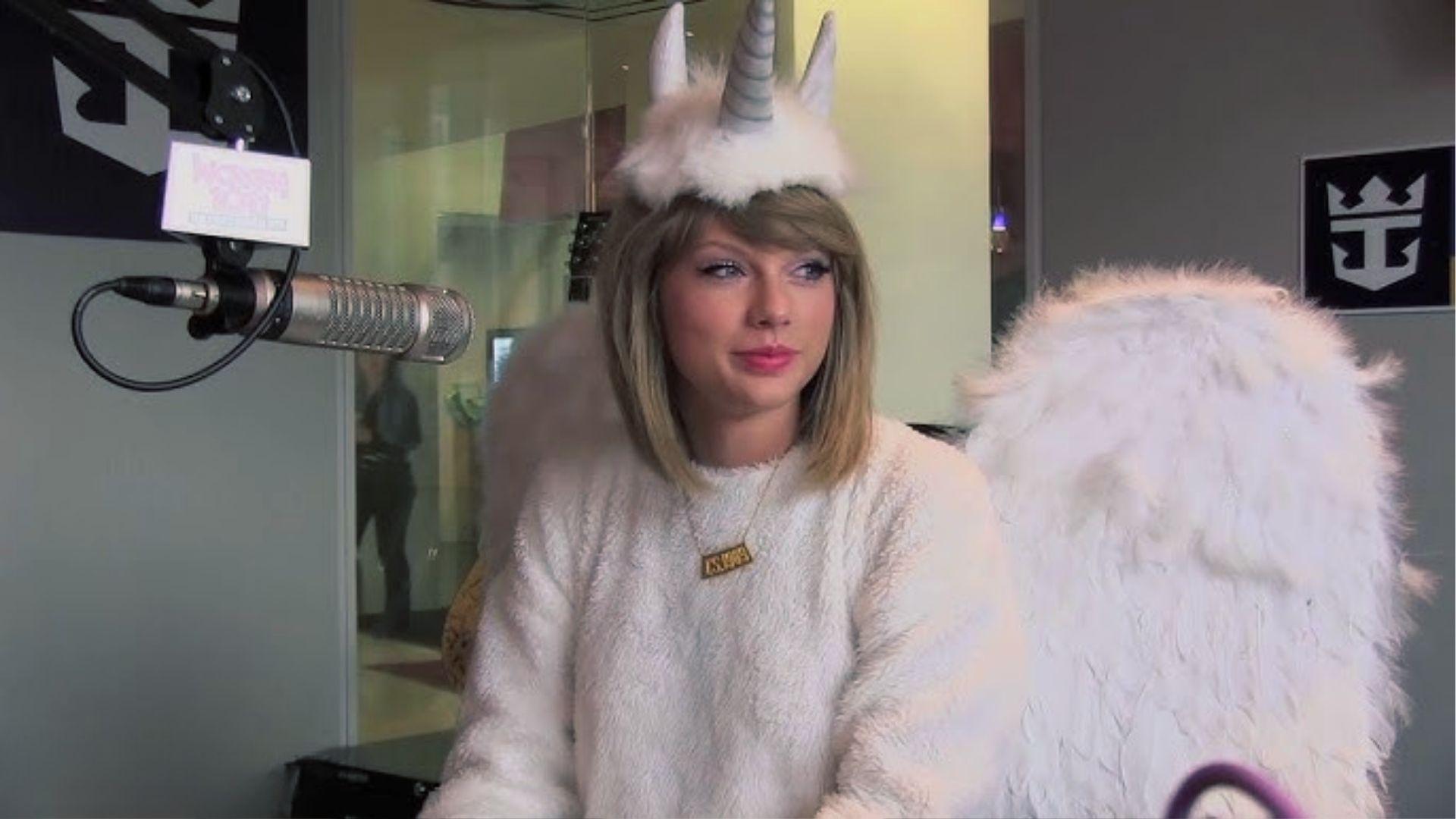 Taylor Swift in white fluffy unicorn costume with horn and wings during radio interview.