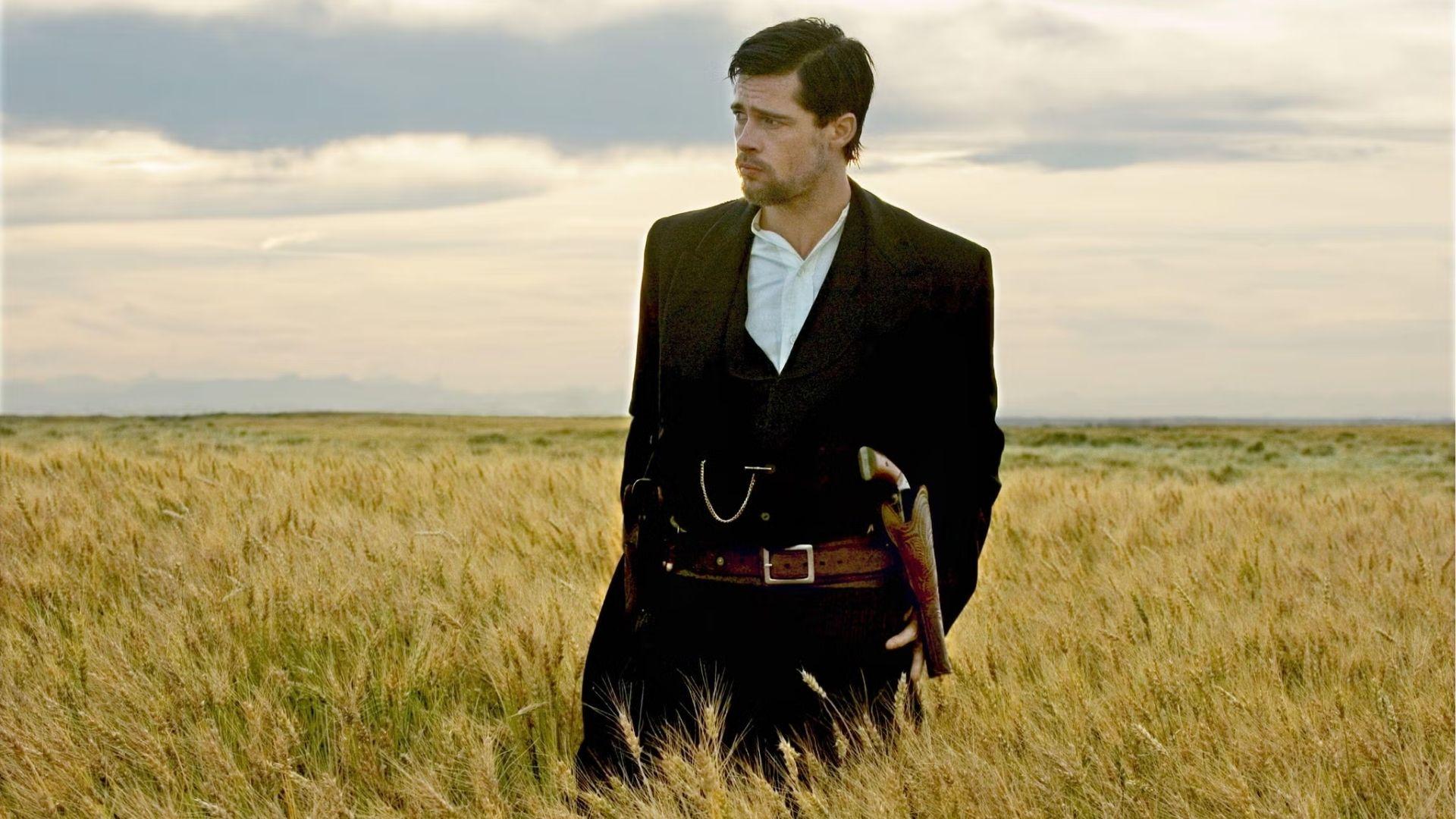 Brad Pitt as Jesse James stands in wheat field wearing black suit, medium hair slicked back