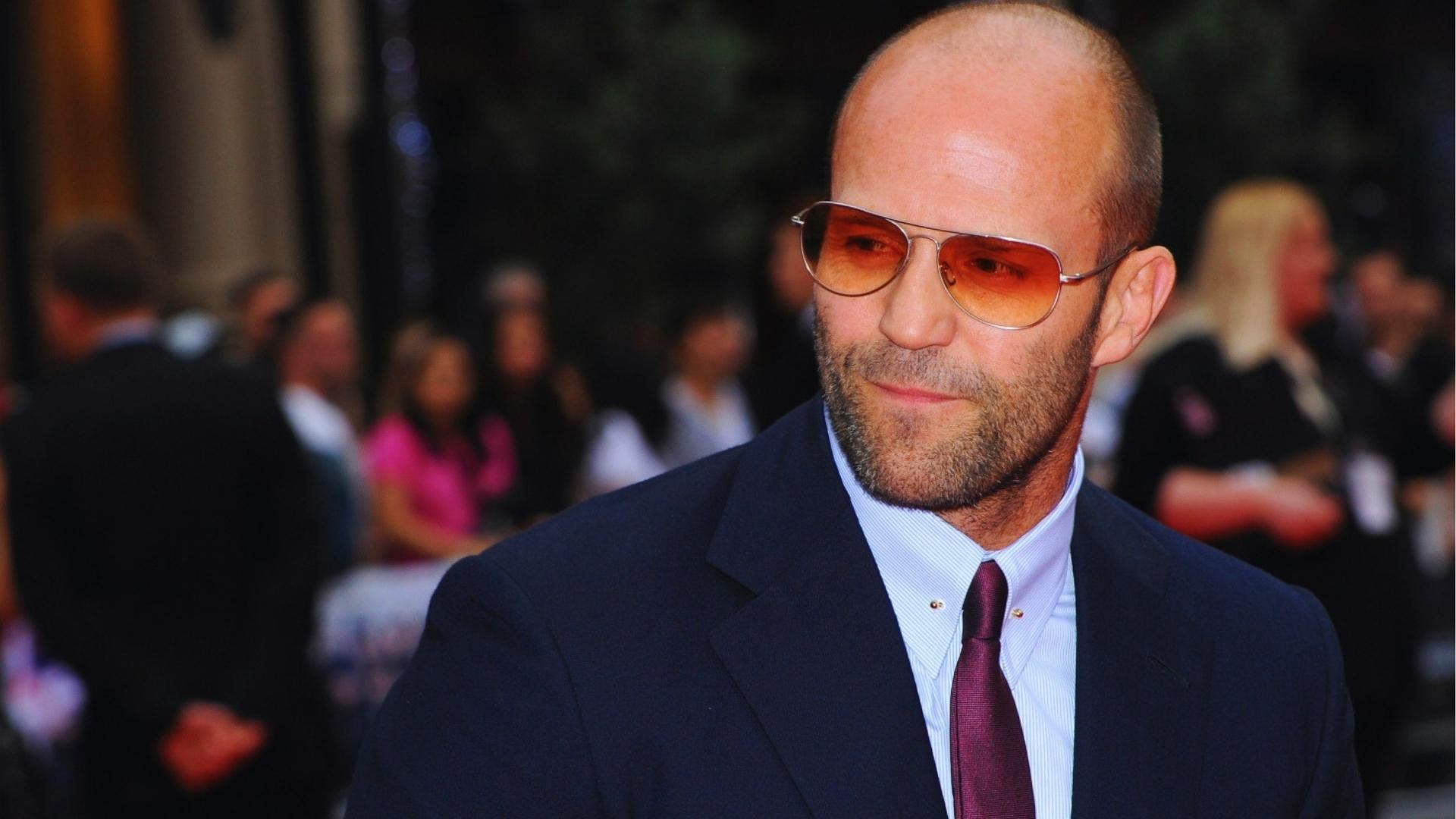 Jason Statham wears tinted aviator sunglasses with a close-shaved head and trimmed beard, showing polished bald style on the red carpet