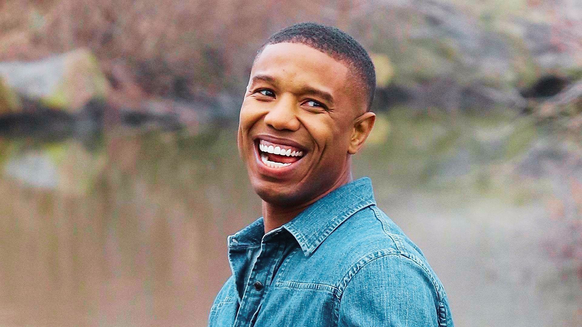 Michael B. Jordan smiling outdoors in a denim shirt, wearing a short crew cut with a natural fade.