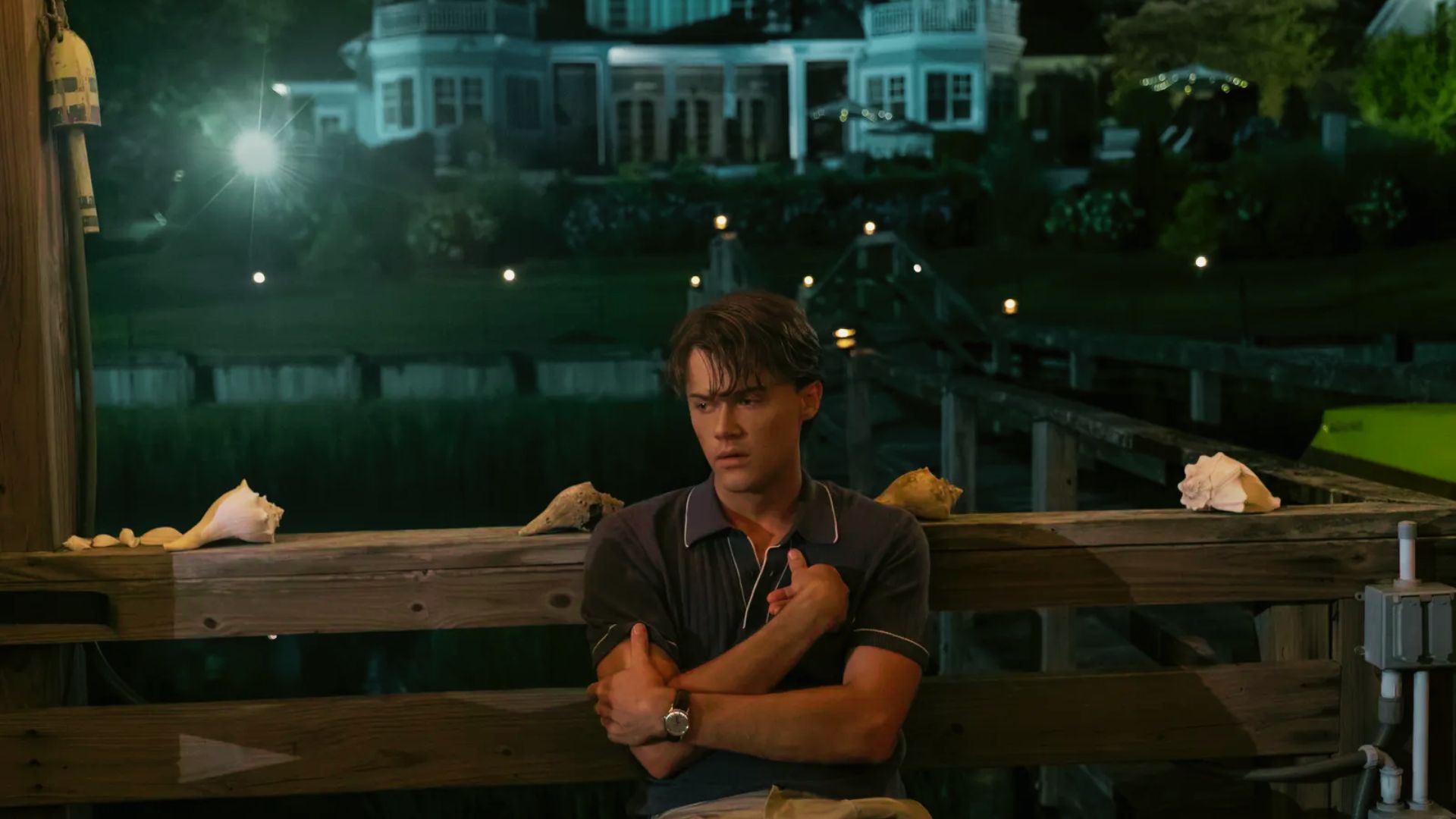 Conrad sits on a dock at night in a navy polo with white piping, arms crossed.