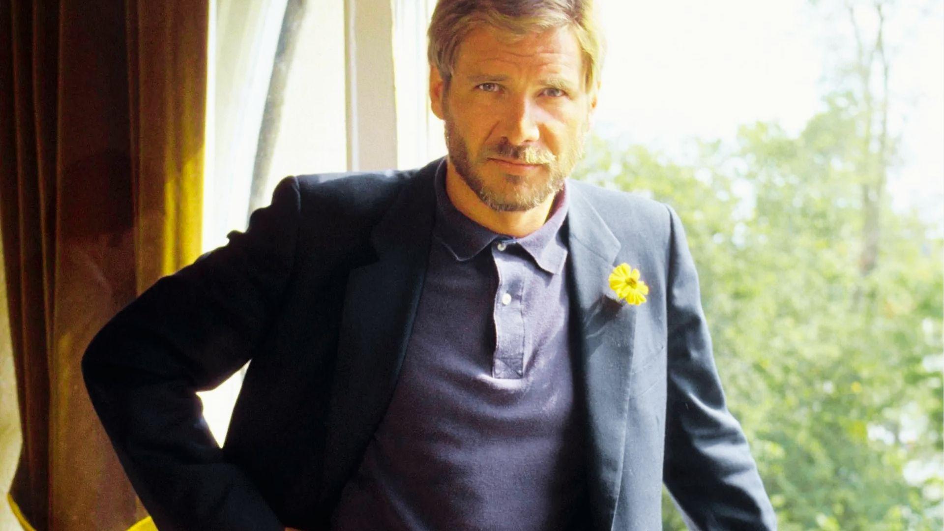 Harrison Ford in dark blazer and polo with yellow flower poses by sunny window
