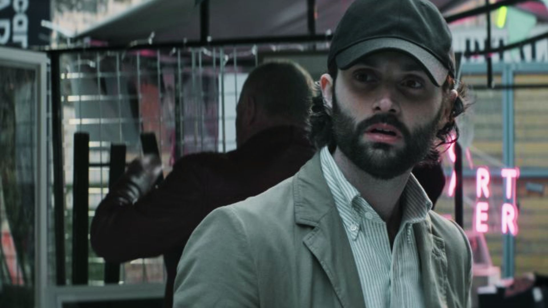 Joe Goldberg wears a black baseball cap and light olive jacket over a striped button-up.