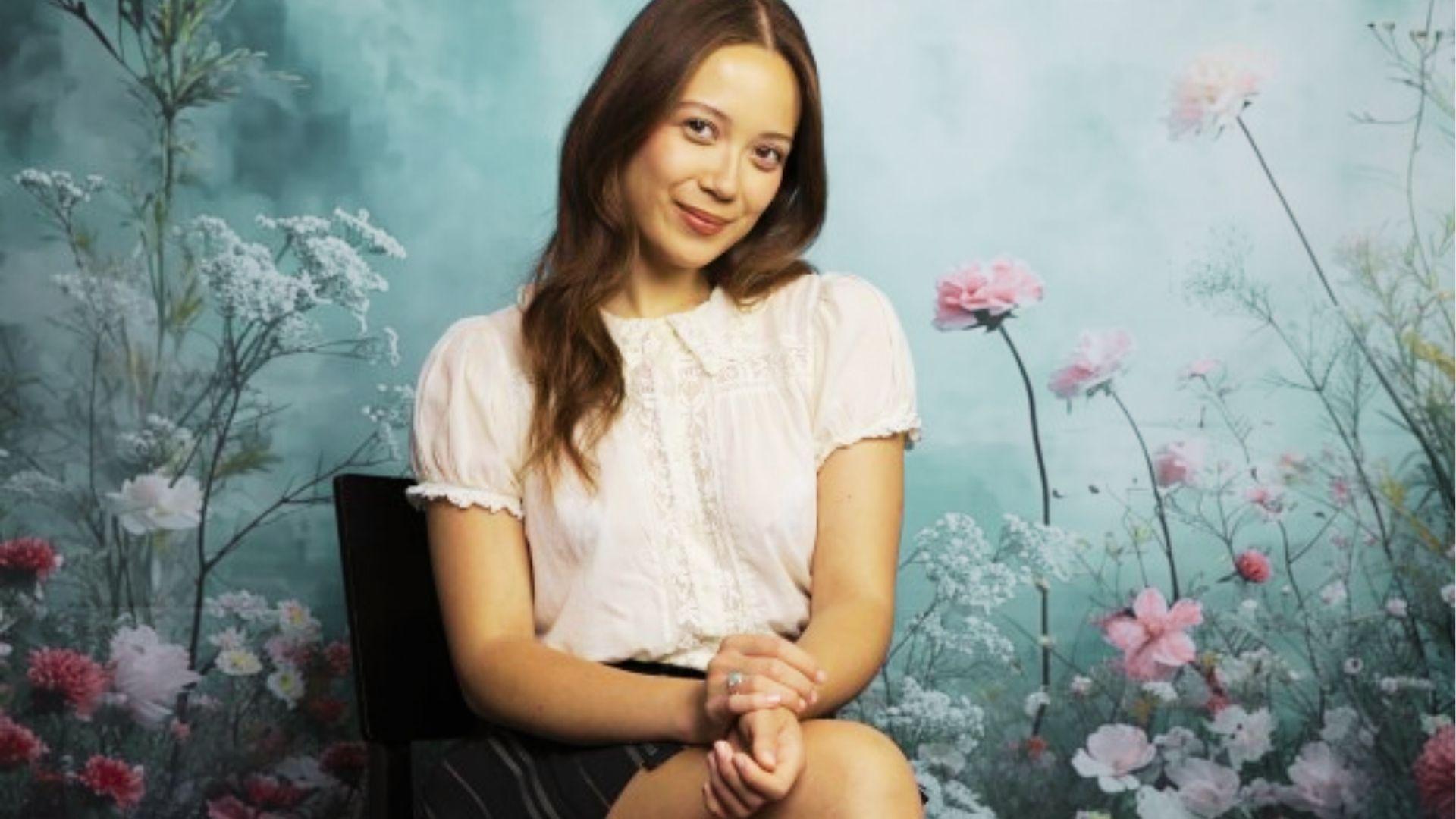 Smiling woman in white lace blouse sits among pastel flowers against soft teal painted background