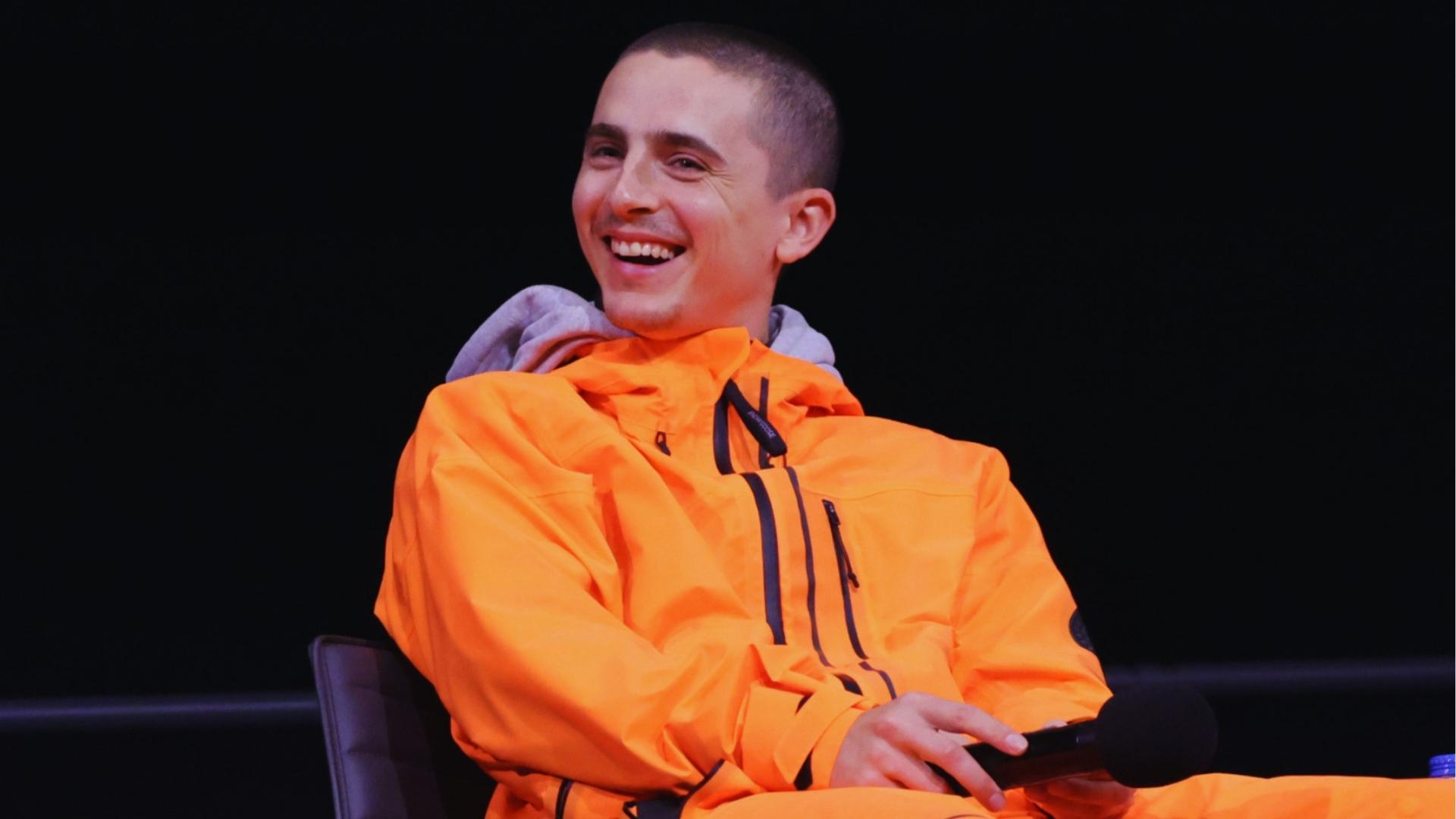 Timothée Chalamet laughs during an interview wearing a bright orange jacket and fresh buzz cut, showing his casual side.