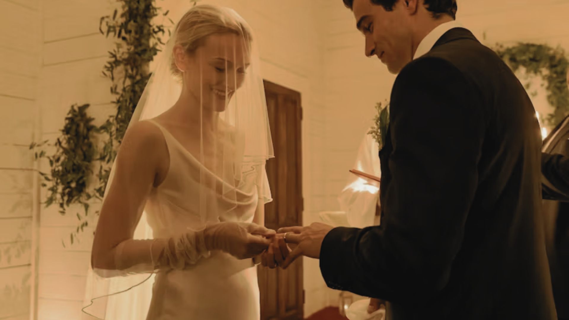 "A bride in a veil smiles gently as she exchanges rings with a groom. The setting features warm lighting and green foliage, creating a romantic ambiance."