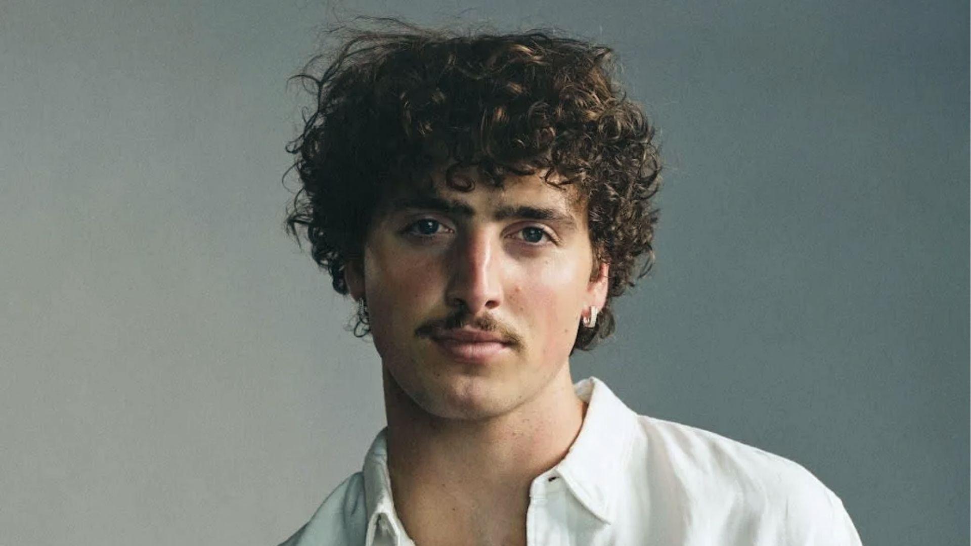 Benson Boone glances aside in white shirt displaying tight voluminous curly hair.