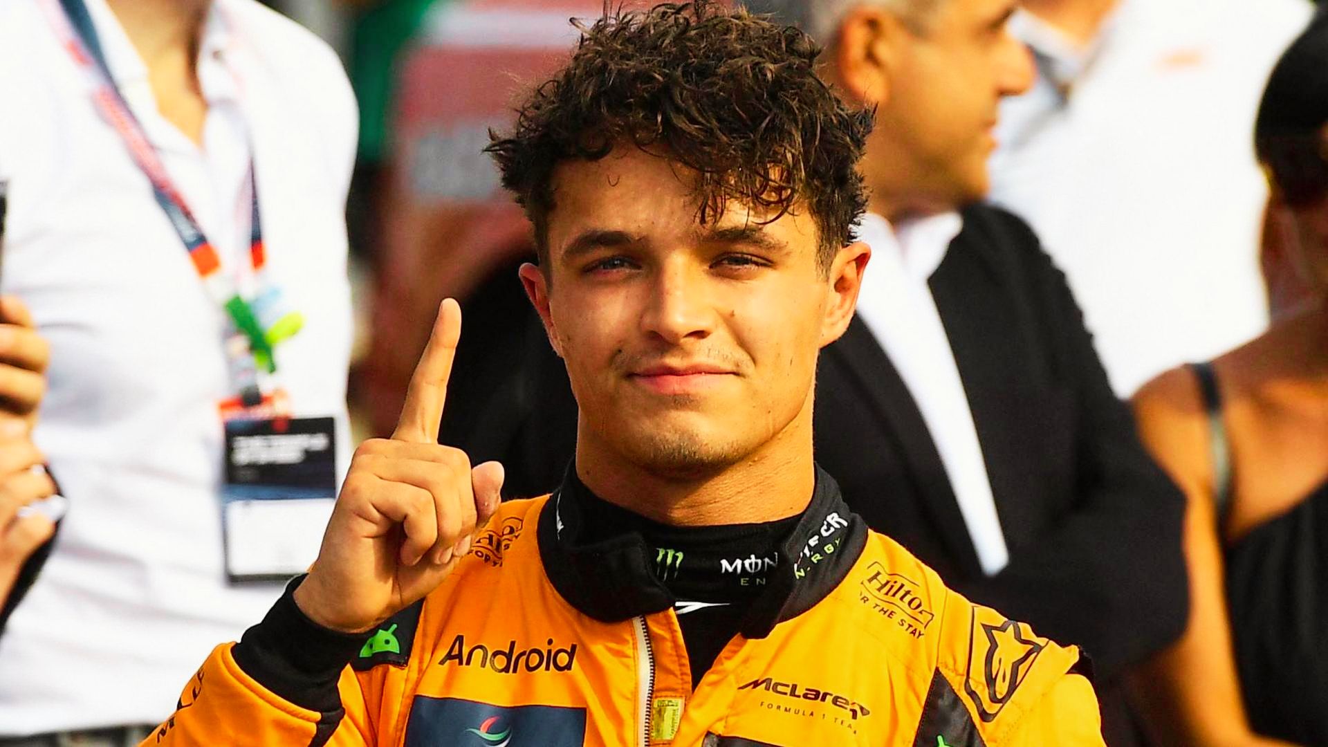 Lando Norris in McLaren racing suit holds up one finger, showing off his tousled curly haircut.