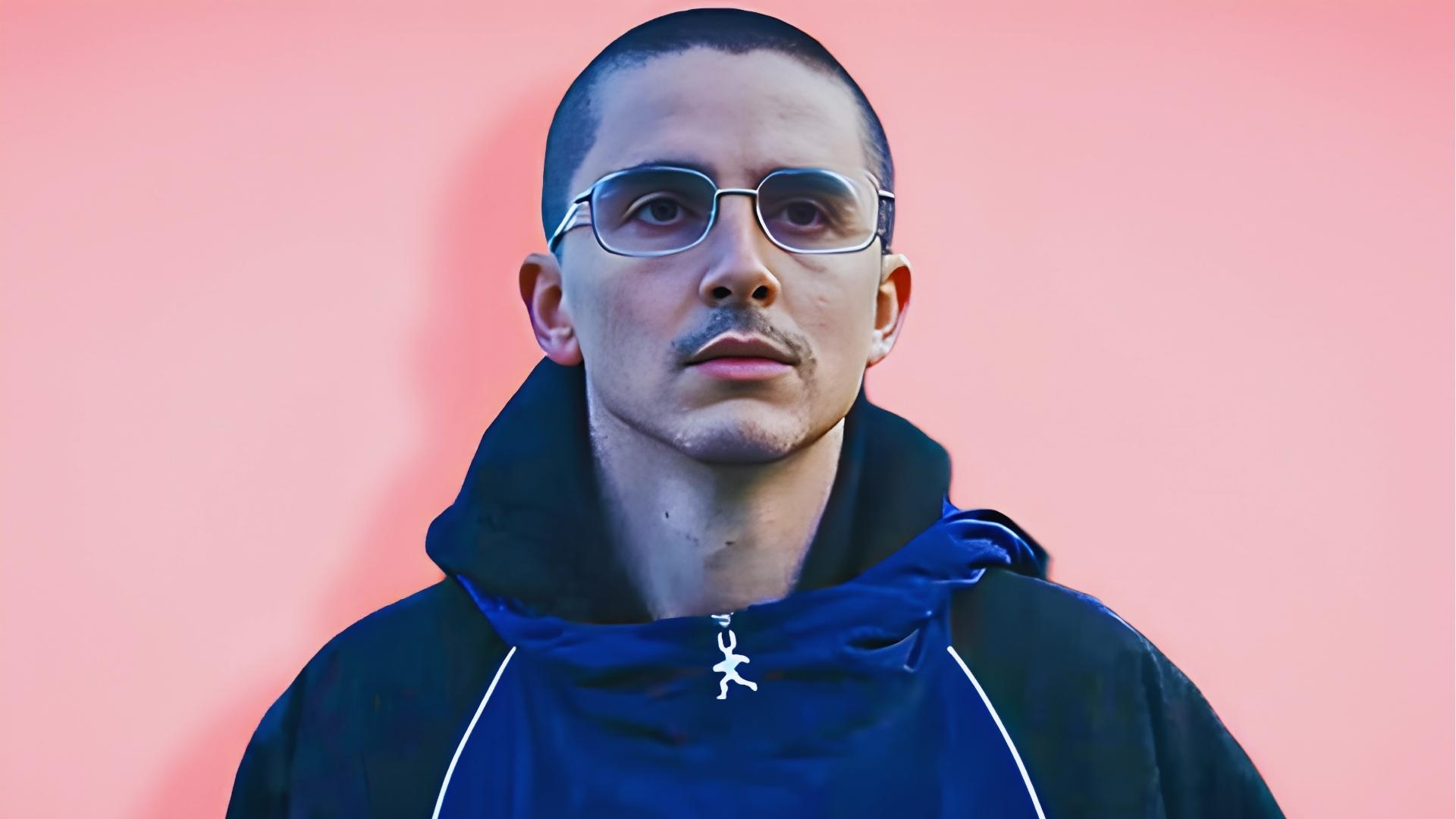 Timothée Chalamet with a close-cropped buzz cut and rectangular glasses poses against a pink background in a navy hoodie.