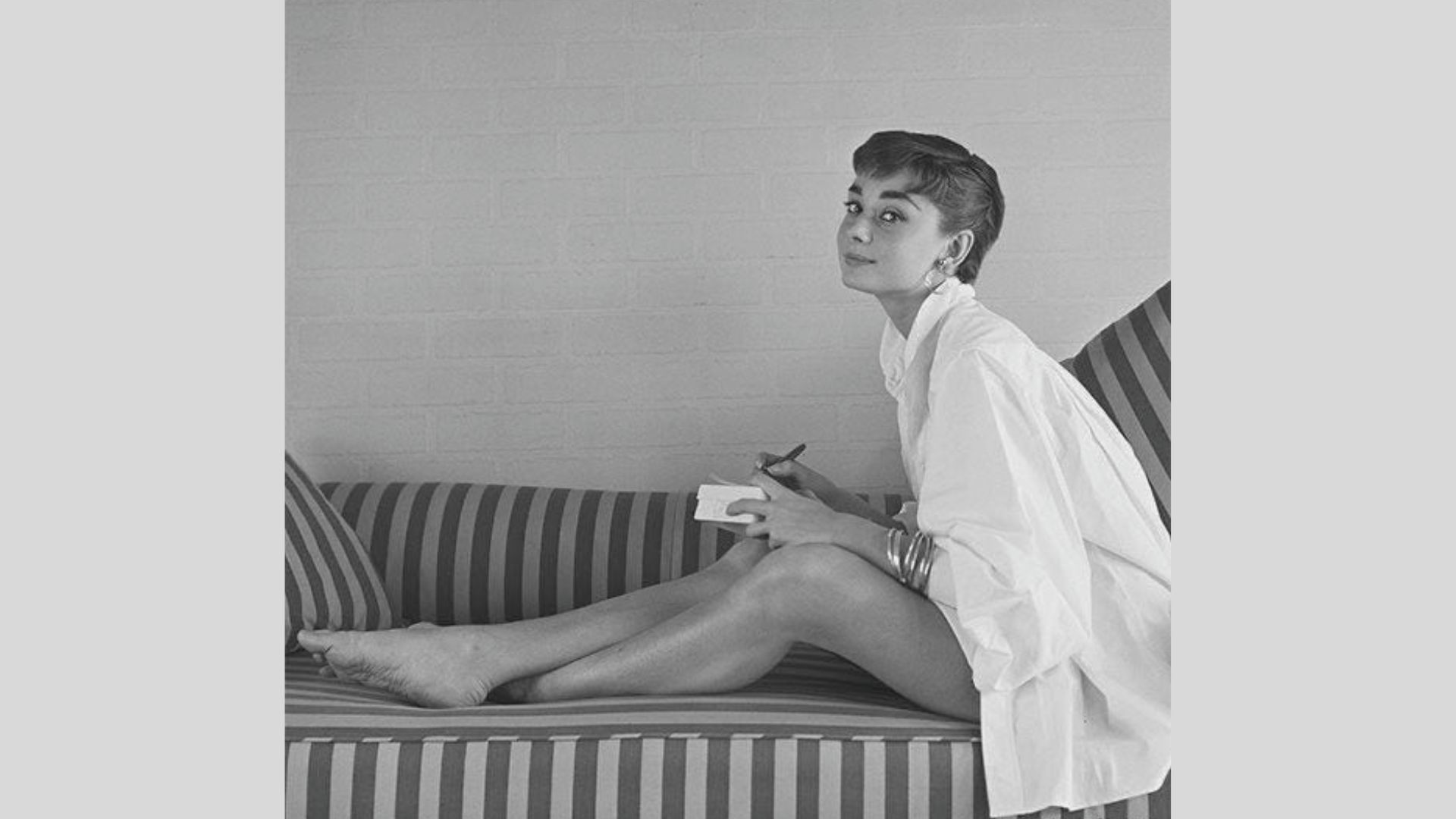 Audrey Hepburn sits barefoot on a striped couch in an oversized white button-down shirt, writing in a notebook.