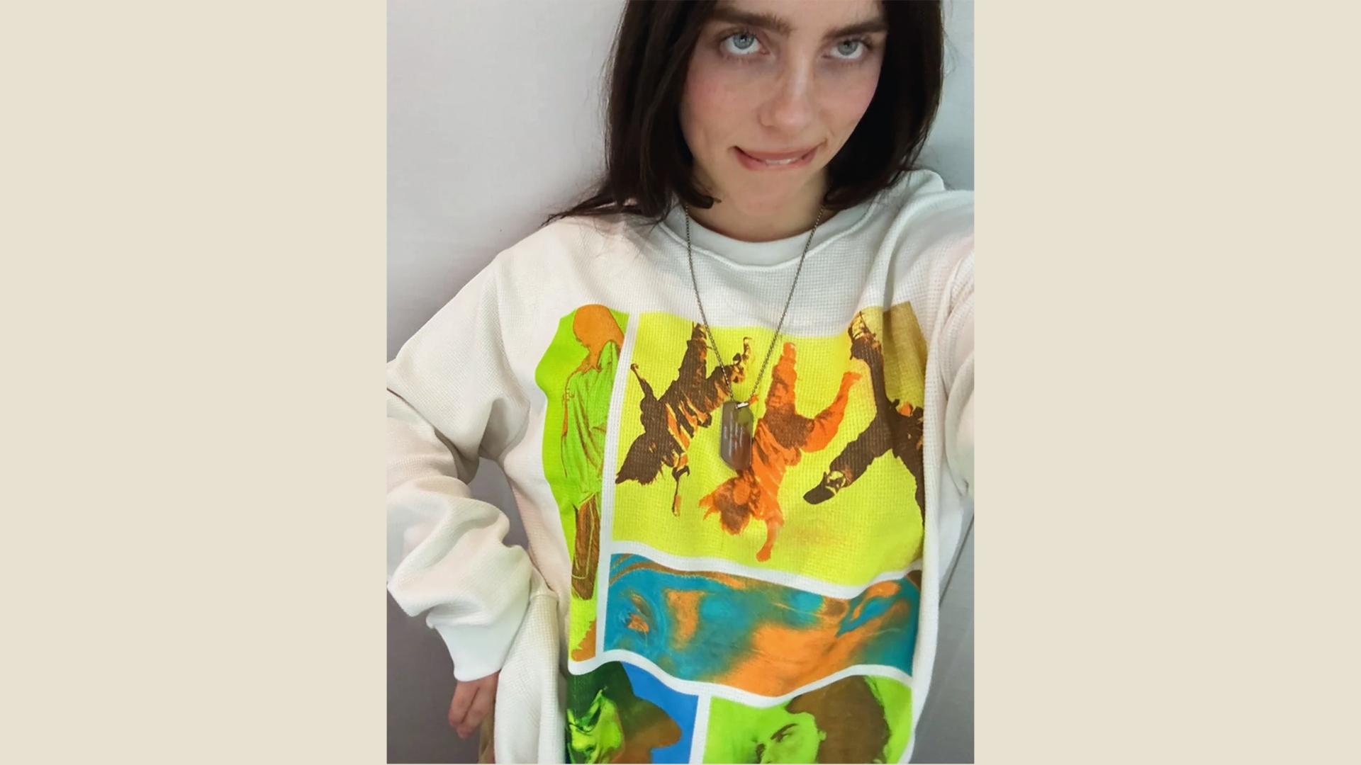 Billie Eilish selfie in oversized white graphic sweatshirt with colorful panels, dog tag necklace, and dark hair.