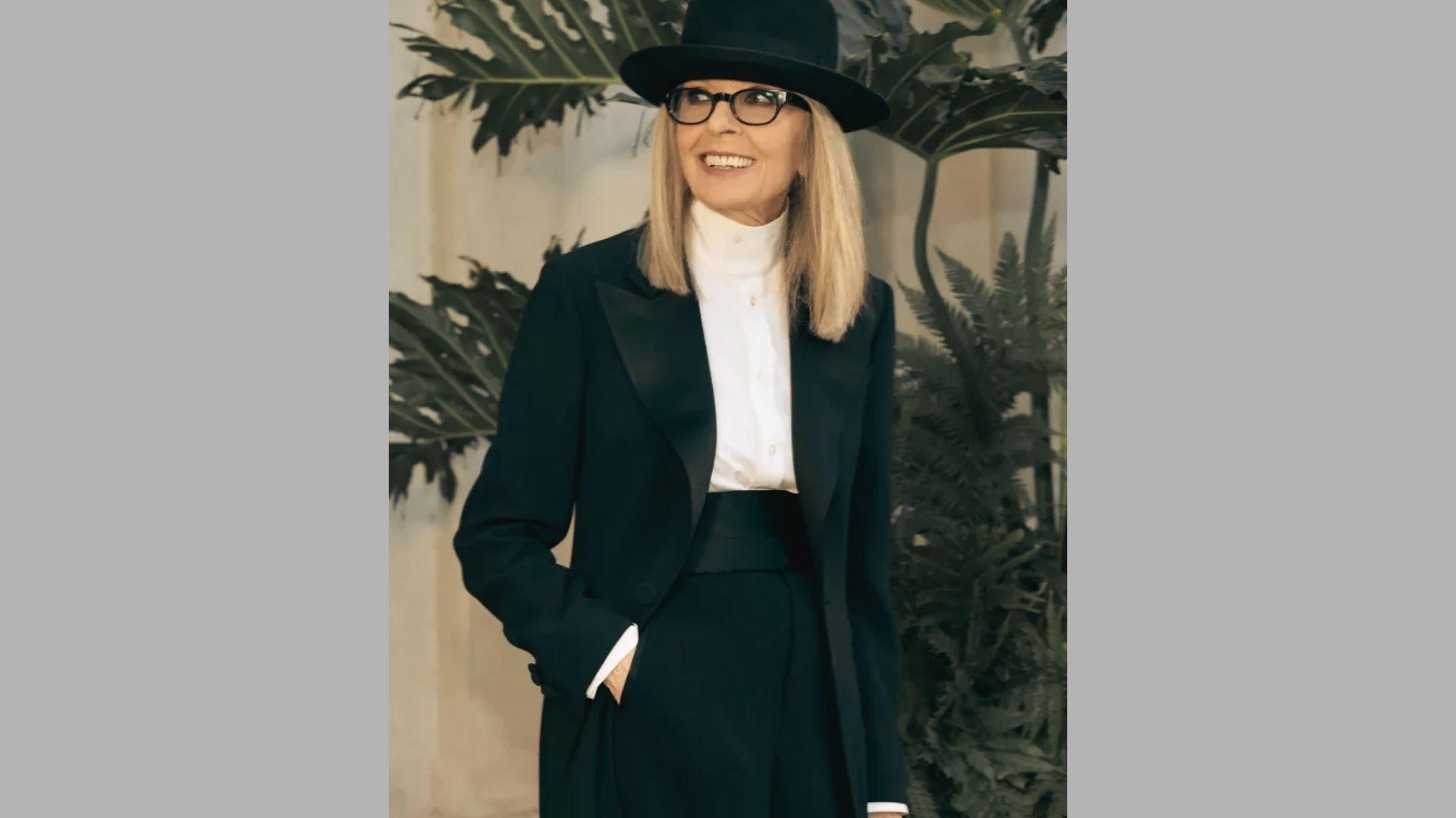 Smiling woman in black tuxedo suit, white shirt, and bowler hat stands before tropical plants