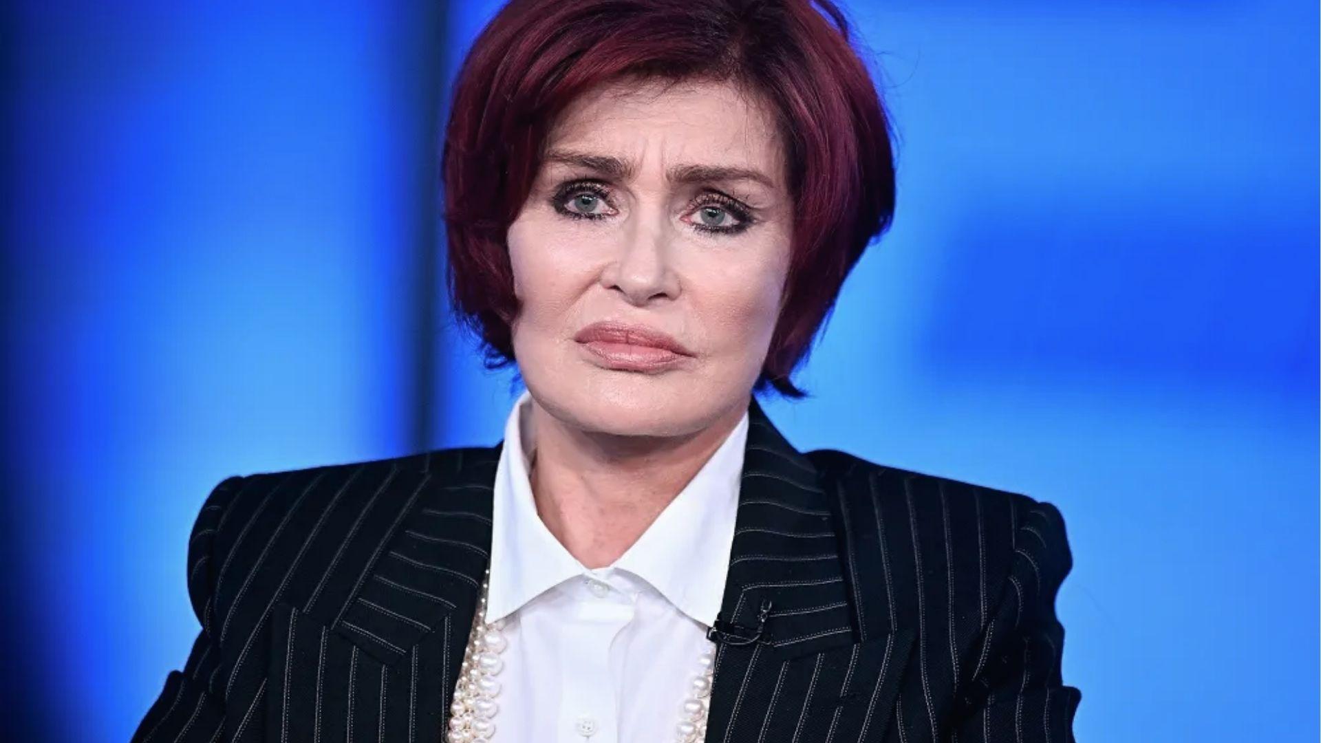 Sharon Osbourne poses in pinstripe blazer, white shirt, and pearl necklace on set