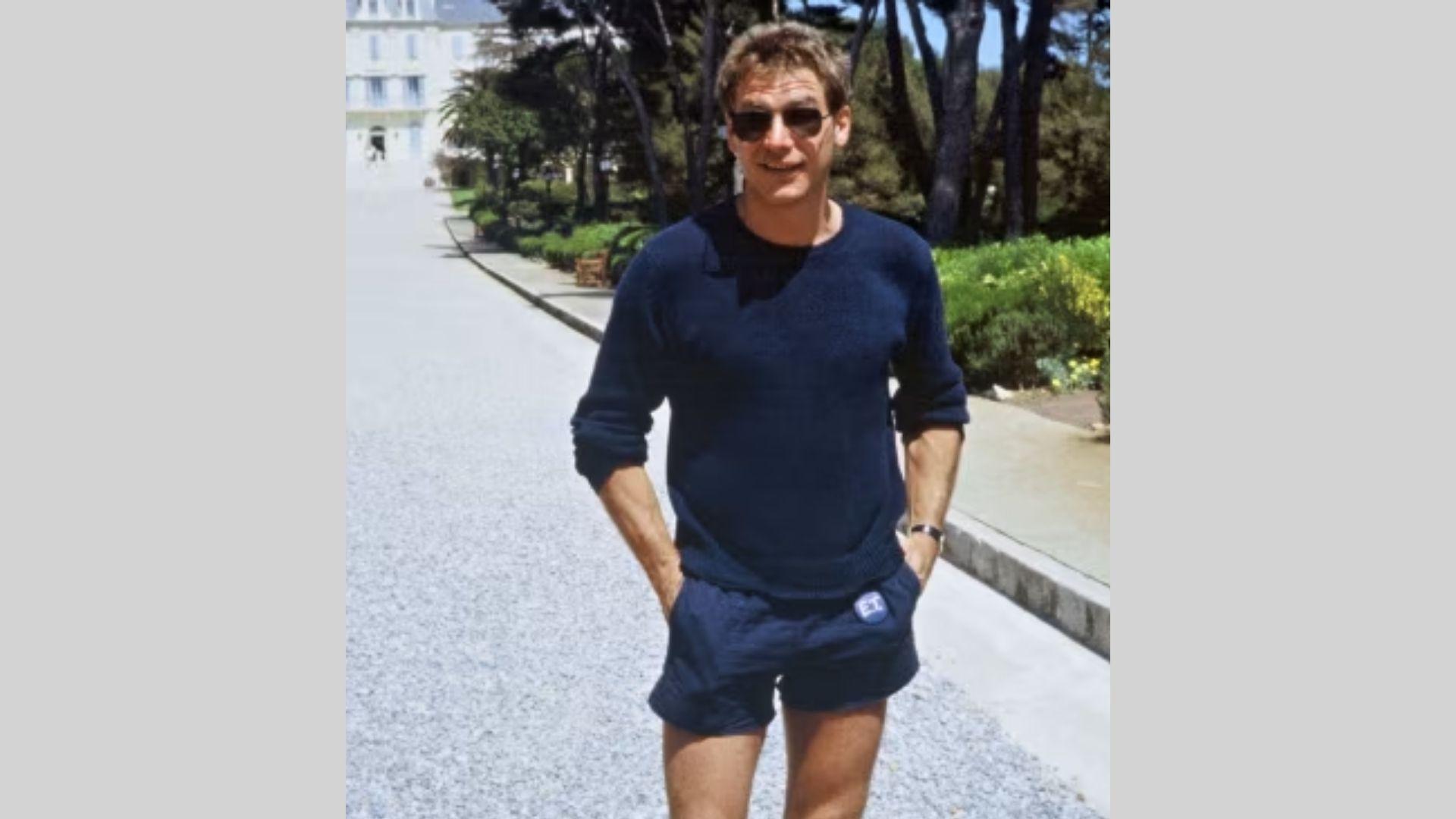 Harrison Ford walks gravel path in navy sweater and shorts with hands in pockets smiling