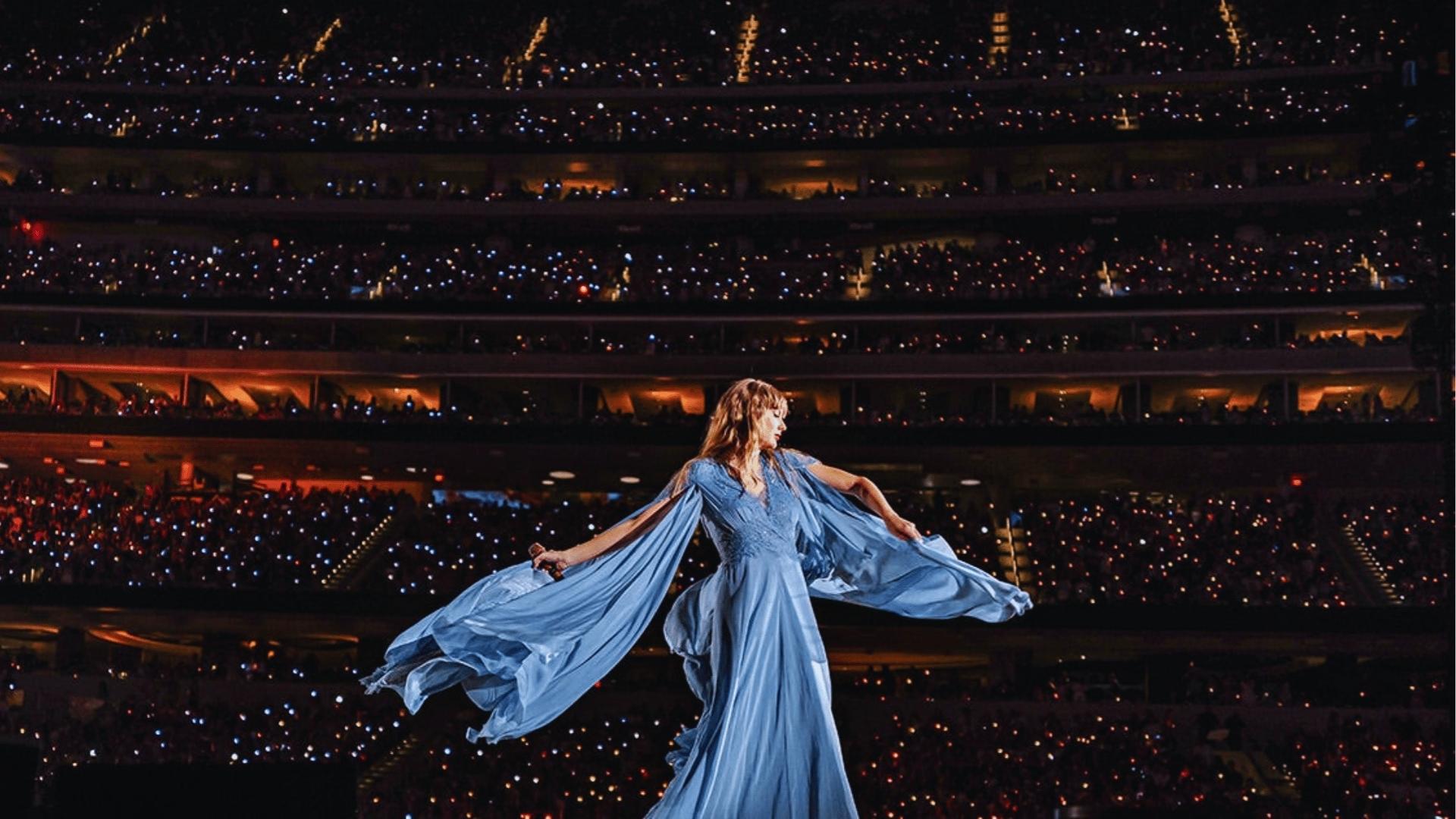 Taylor Swift twirls on stage in a flowing sky-blue folklore gown with cape sleeves during Eras Tour stadium show.